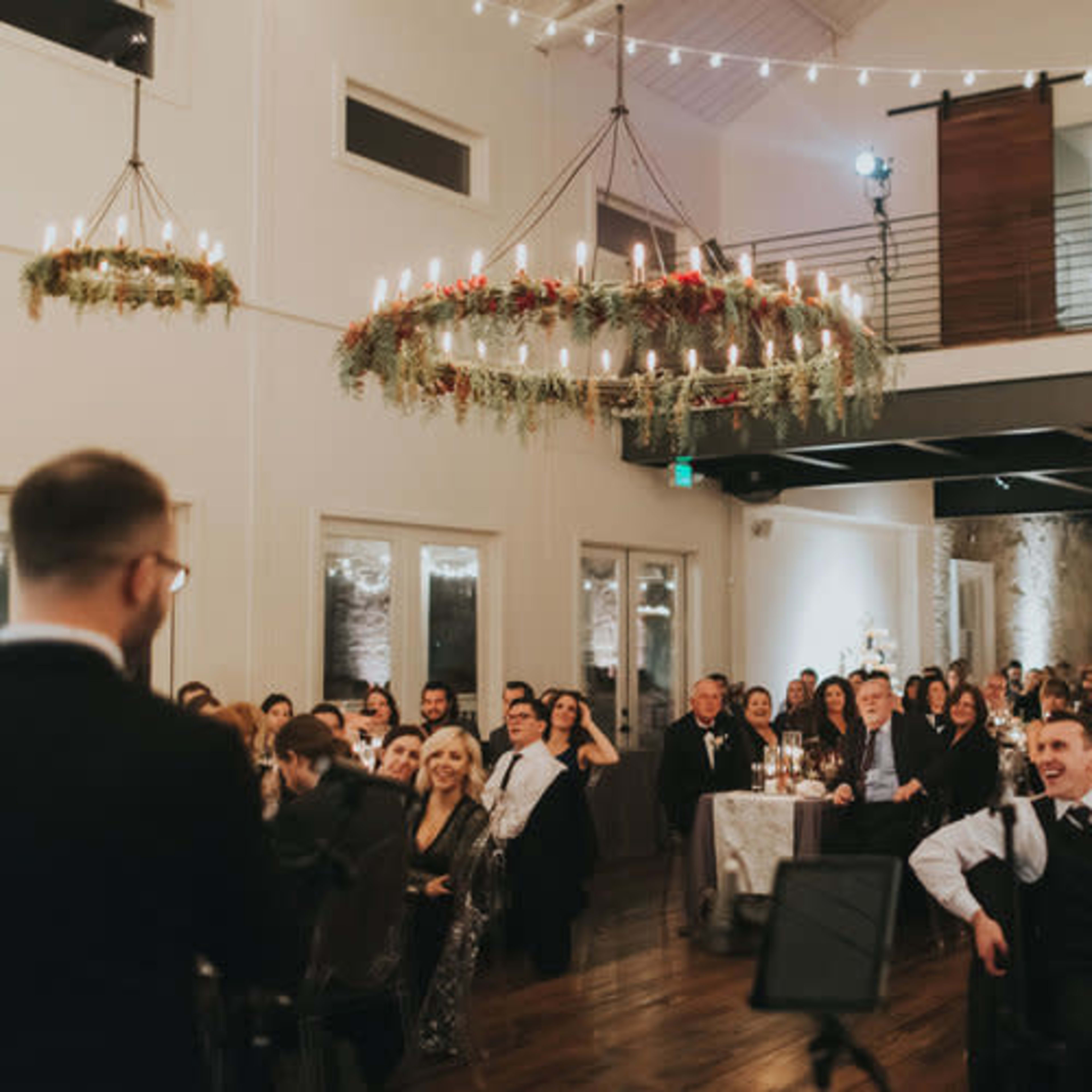 A crowd of guests dressed in formal attire attentively listens to a speaker at an event in a well-lit venue with chandeliers and elegant decor.