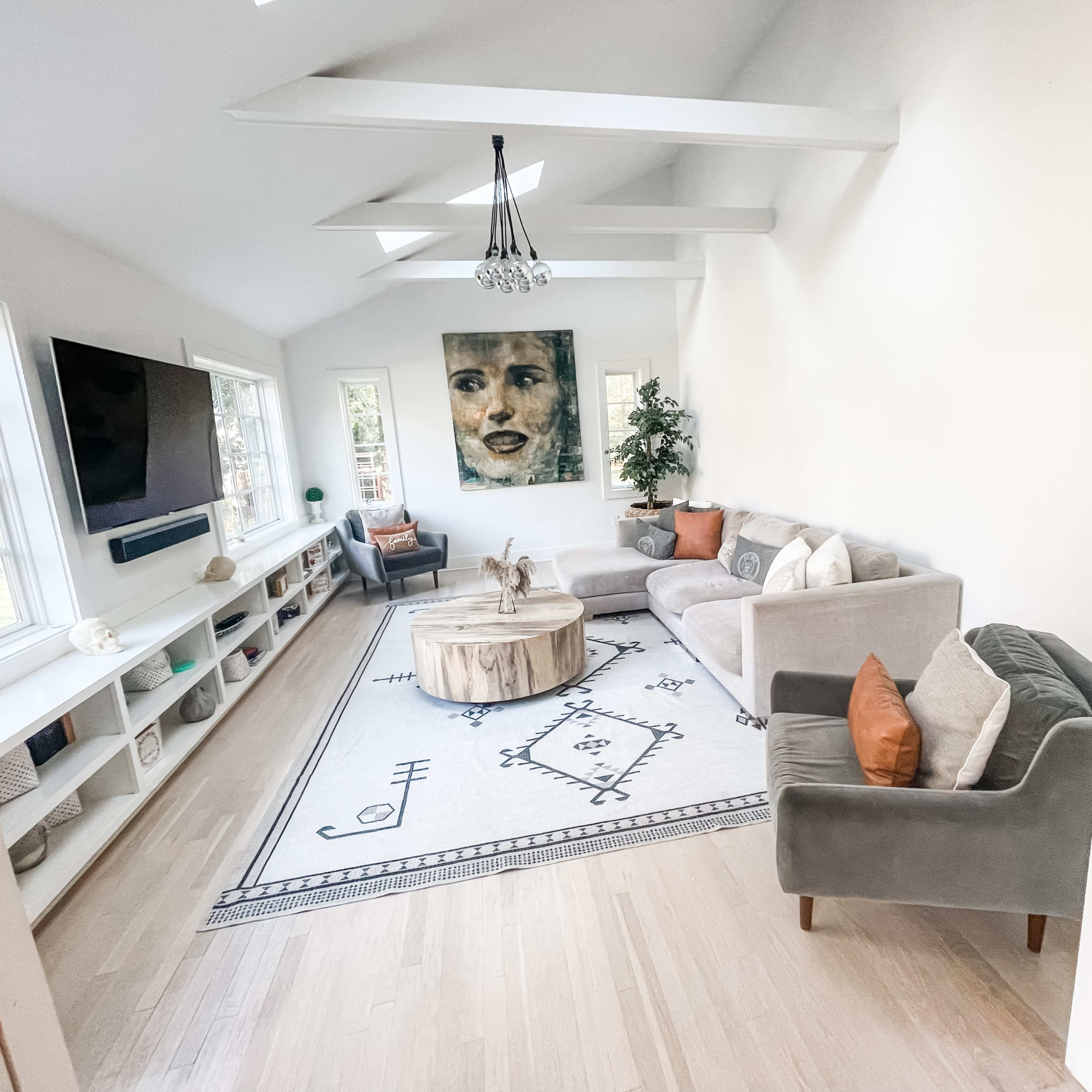 A modern living room with a gray sofa and a green armchair, a round wooden coffee table, and a large artwork on the wall, with a light-colored rug and a television mounted on a white cabinet.