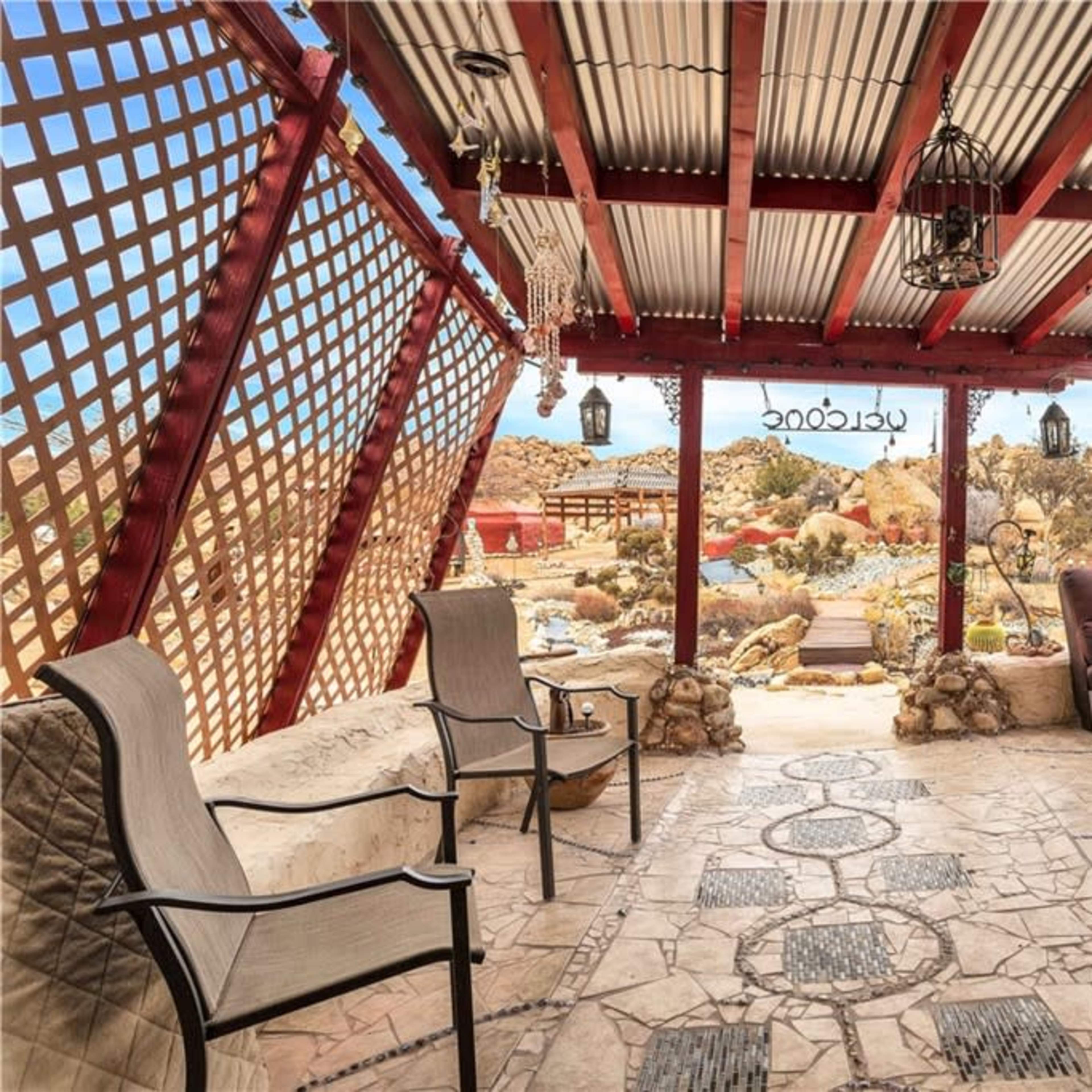 A covered outdoor patio with stone flooring, a lattice structure, and comfortable seating overlooking a desert landscape.