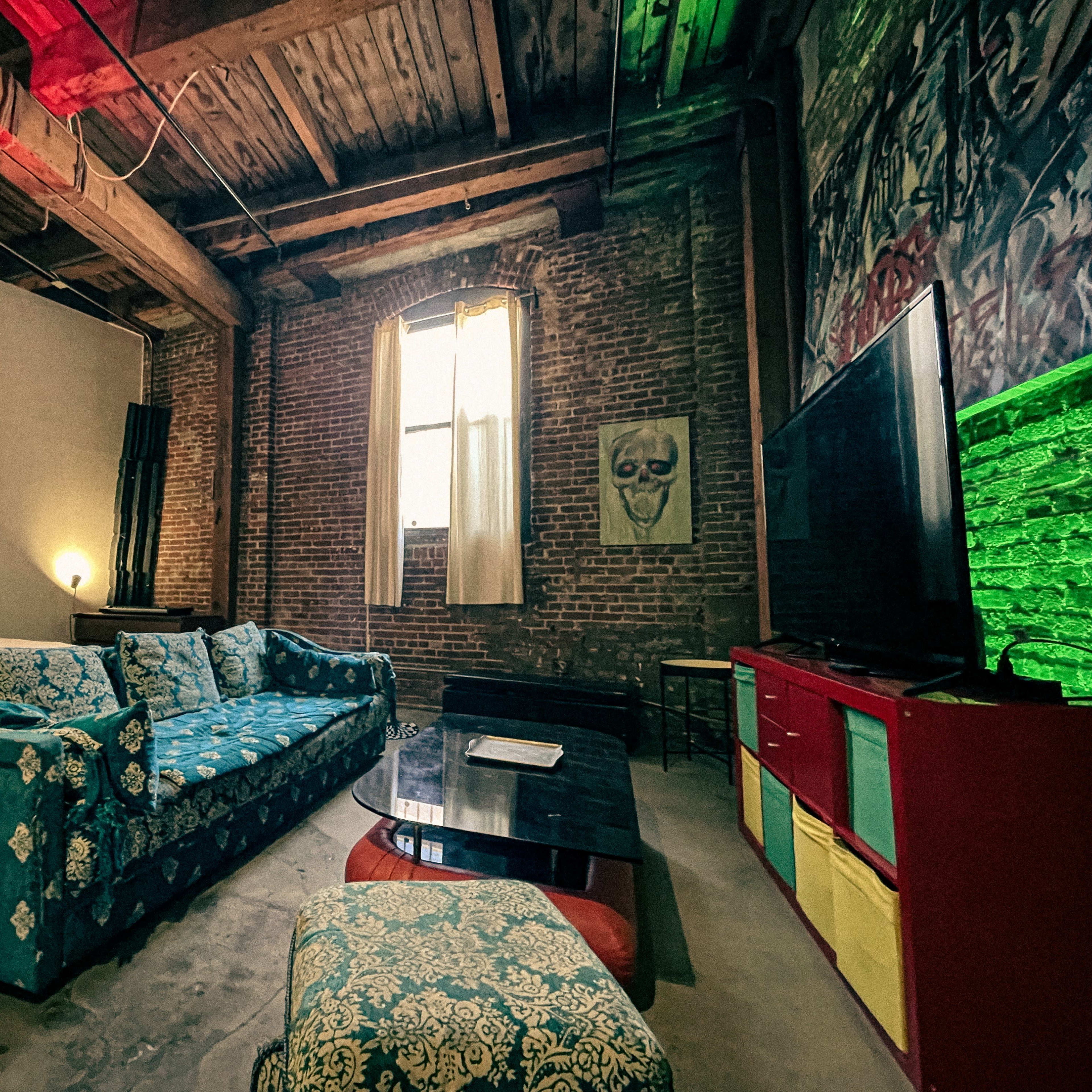 A spacious loft-style living area featuring exposed brick walls, a colorful couch, a coffee table, and a large window with sheer curtains.