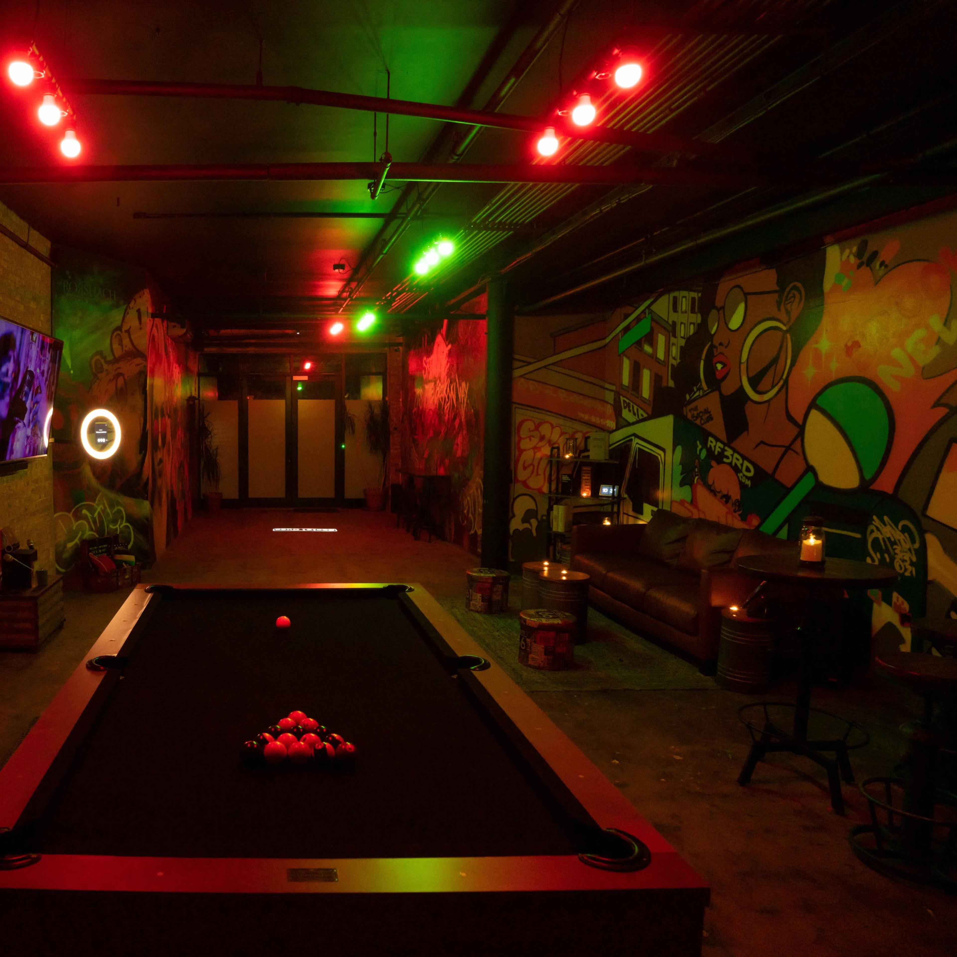 A dimly lit lounge area featuring a pool table, illuminated by red and green lights, with vibrant murals on the walls and a television on one side.