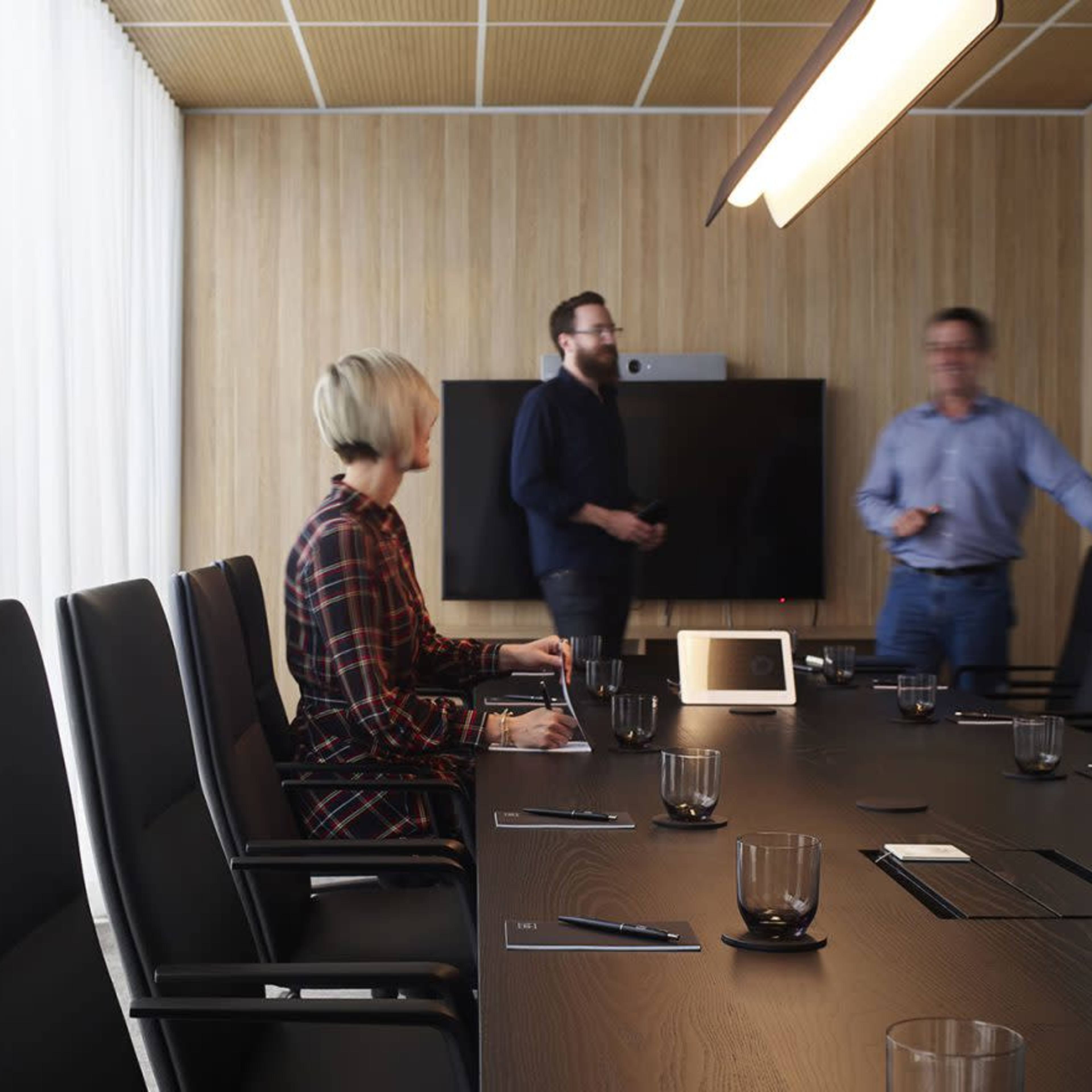 A modern conference room features a long table with black chairs, while three individuals are gathered near a presentation screen.