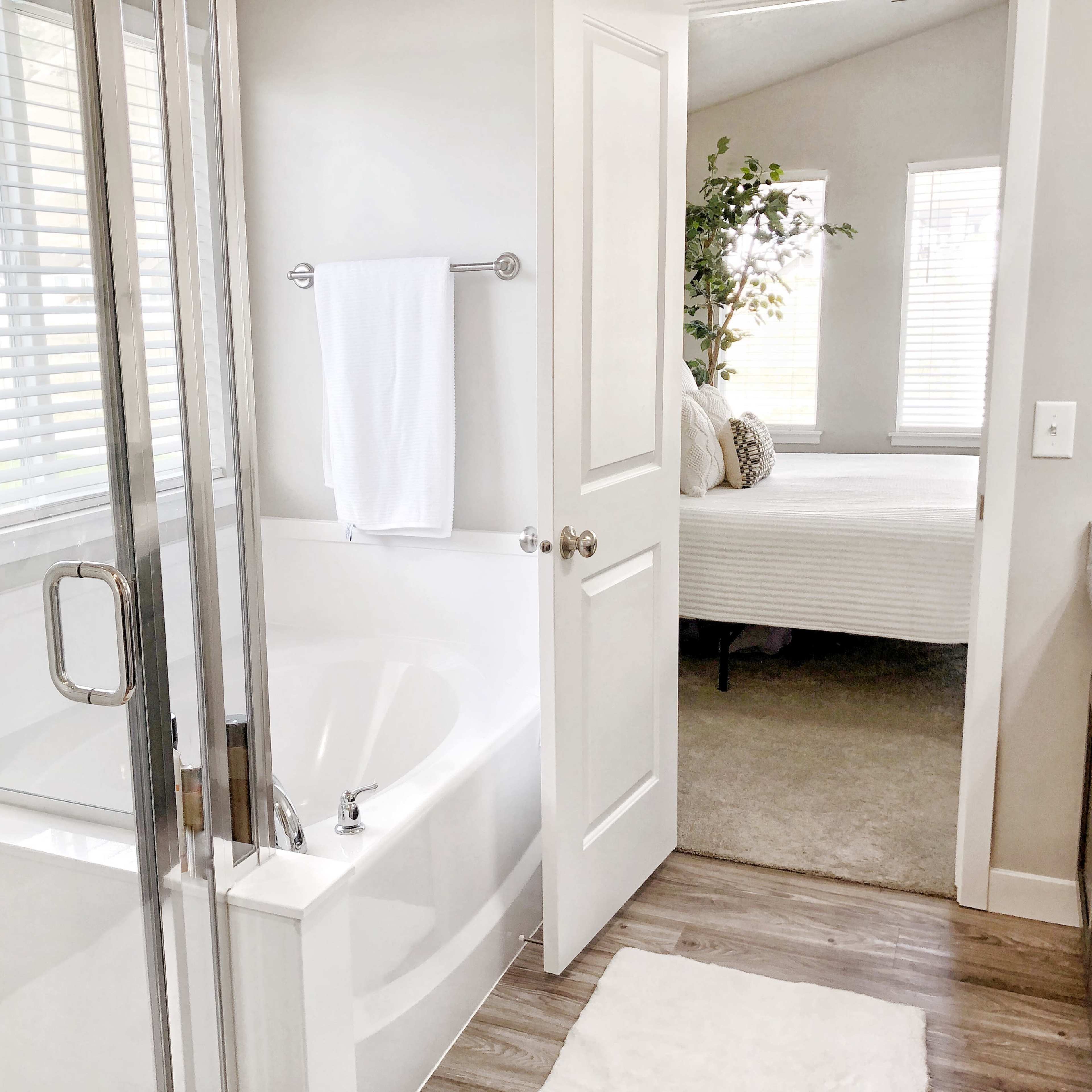 A bright bathroom featuring a glass shower, a soaking tub, and a doorway leading to a carpeted bedroom with large windows.