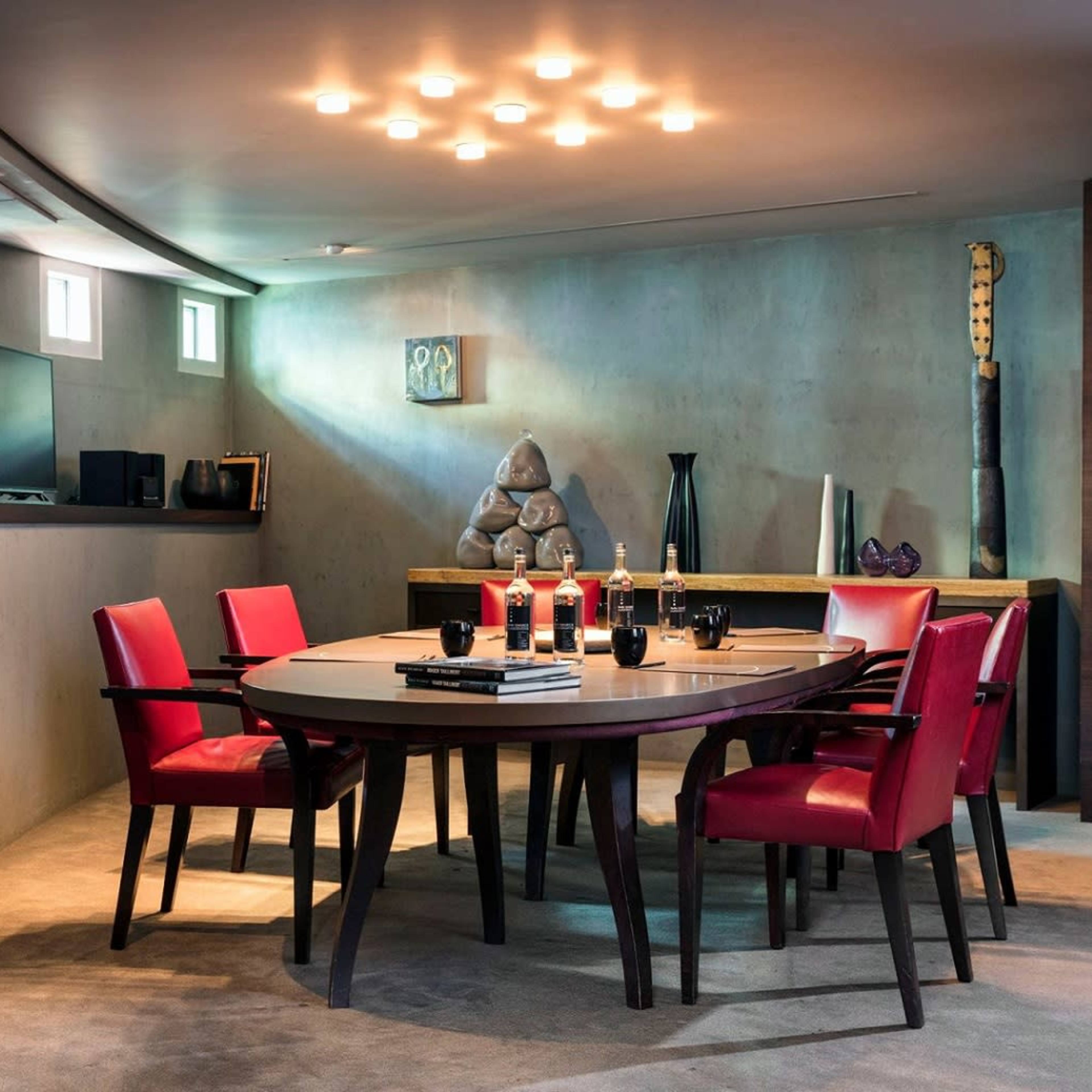 A modern dining room features a round table surrounded by red chairs, with a concrete wall and artistic decor in the background.