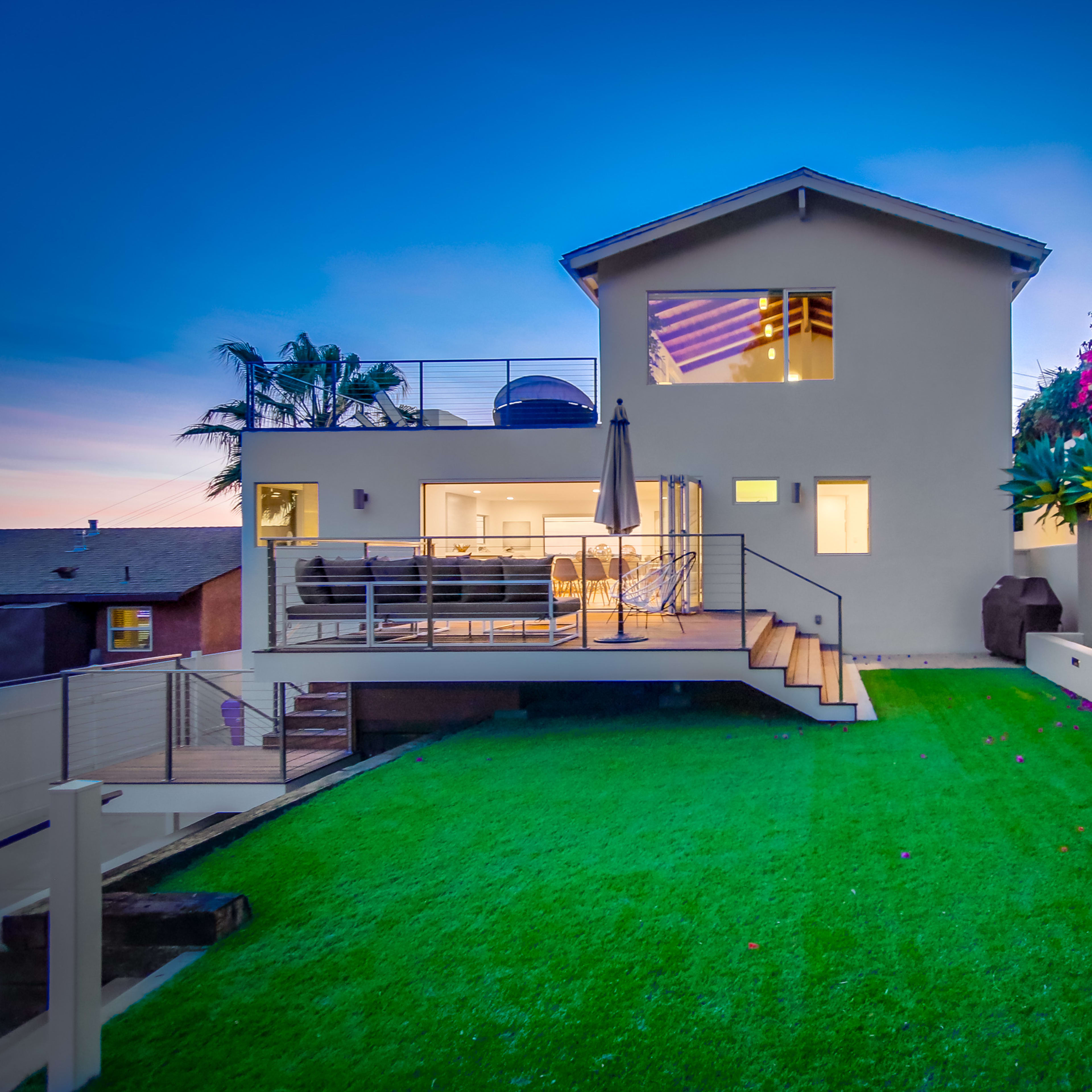 A modern two-story house with a patio and green lawn is set against a backdrop of colorful foliage during twilight.