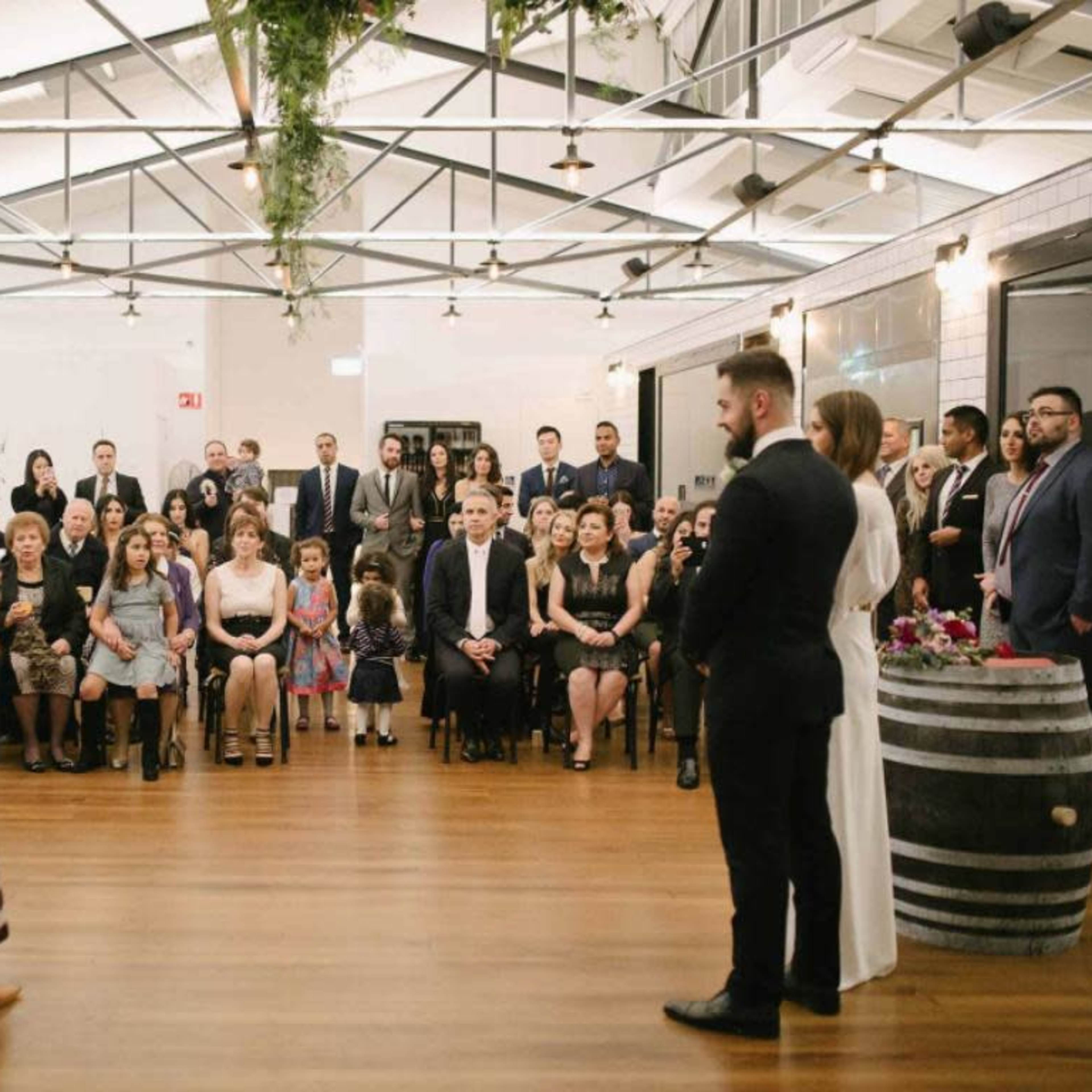A crowd of guests sits on chairs in a venue as a person in traditional attire stands before a couple exchanging vows.