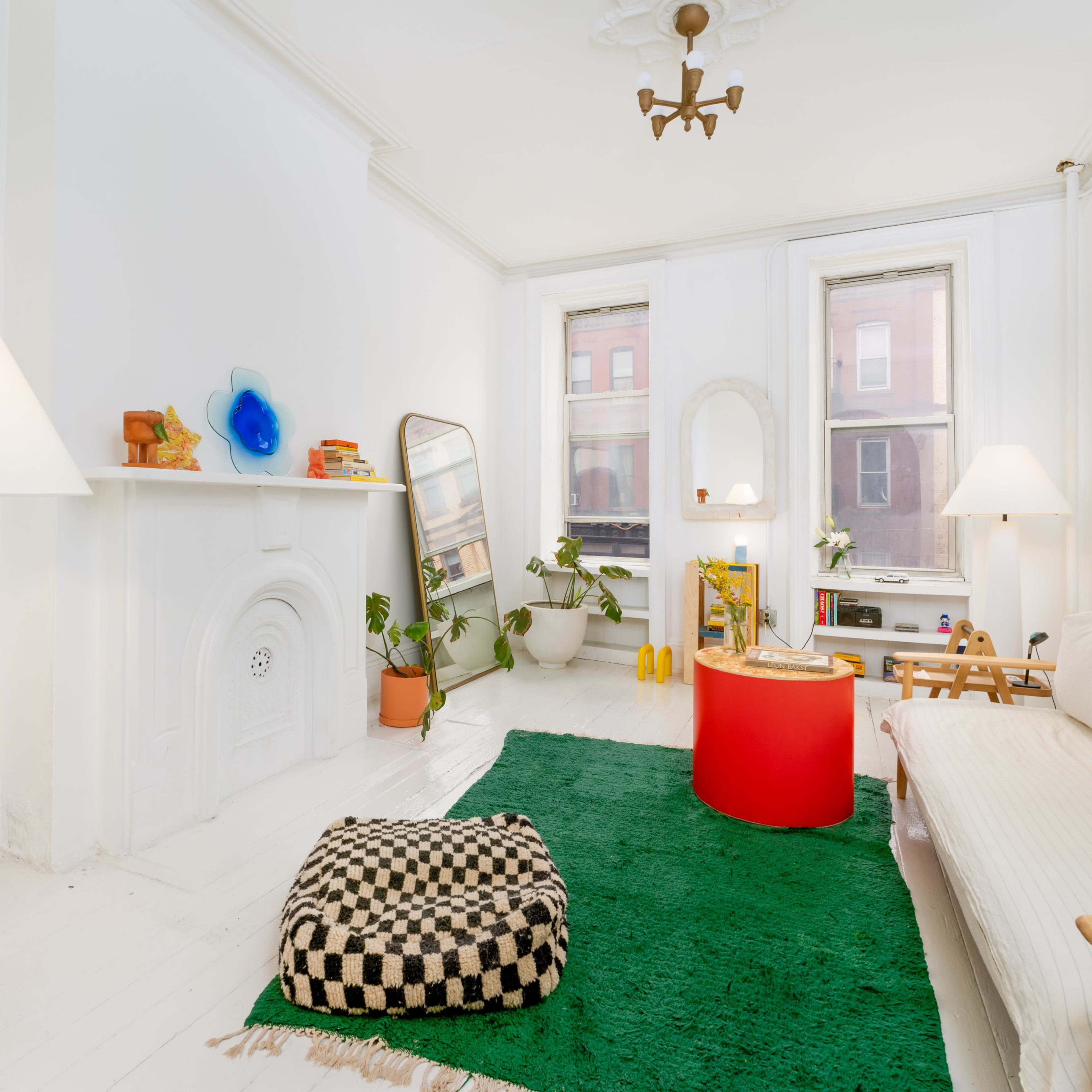 A bright and minimalist living room with a white sofa, a green rug, a round red table, and large windows allowing natural light.