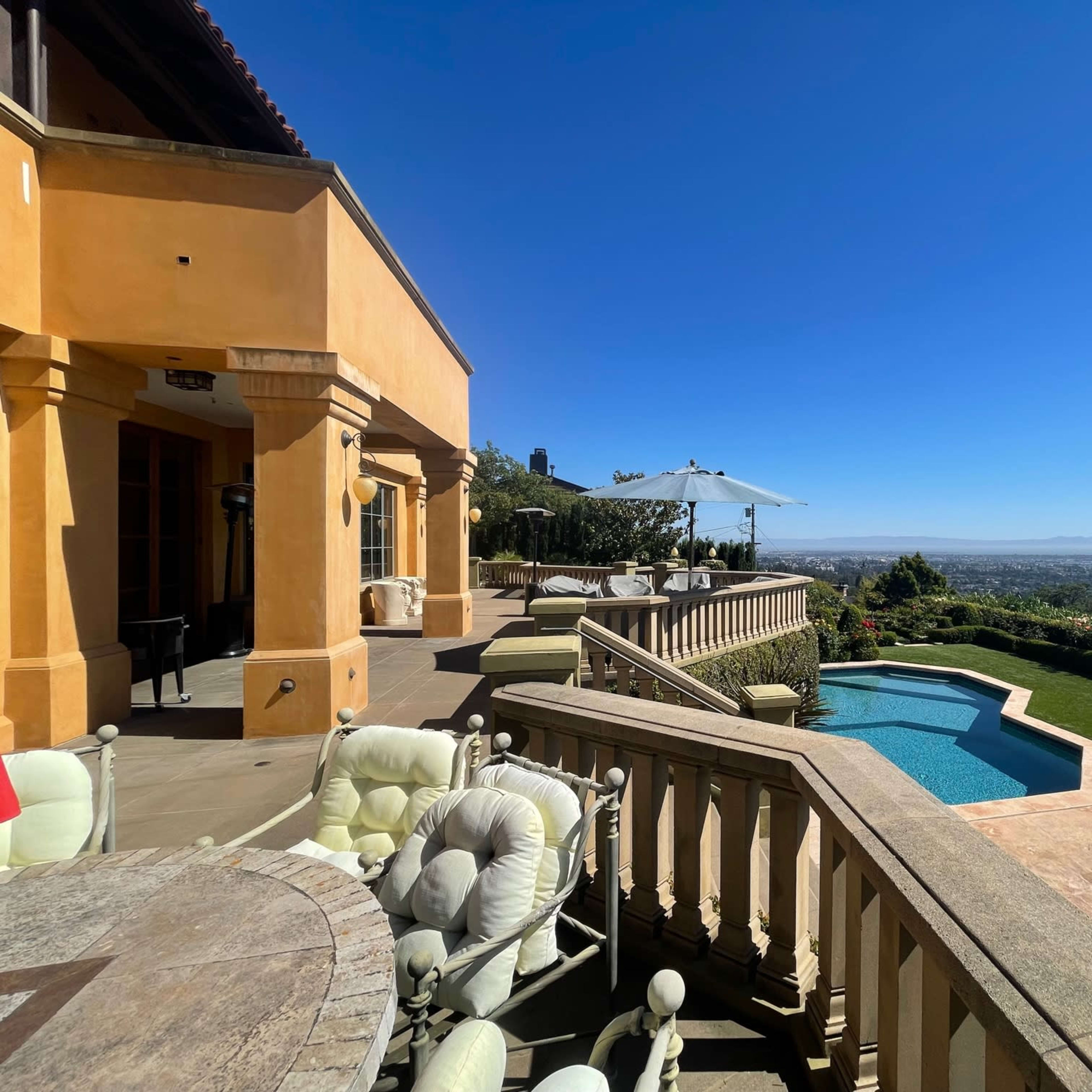 A spacious terrace with outdoor seating surrounds a swimming pool, overlooking a panoramic view of a valley and distant hills under a clear blue sky.