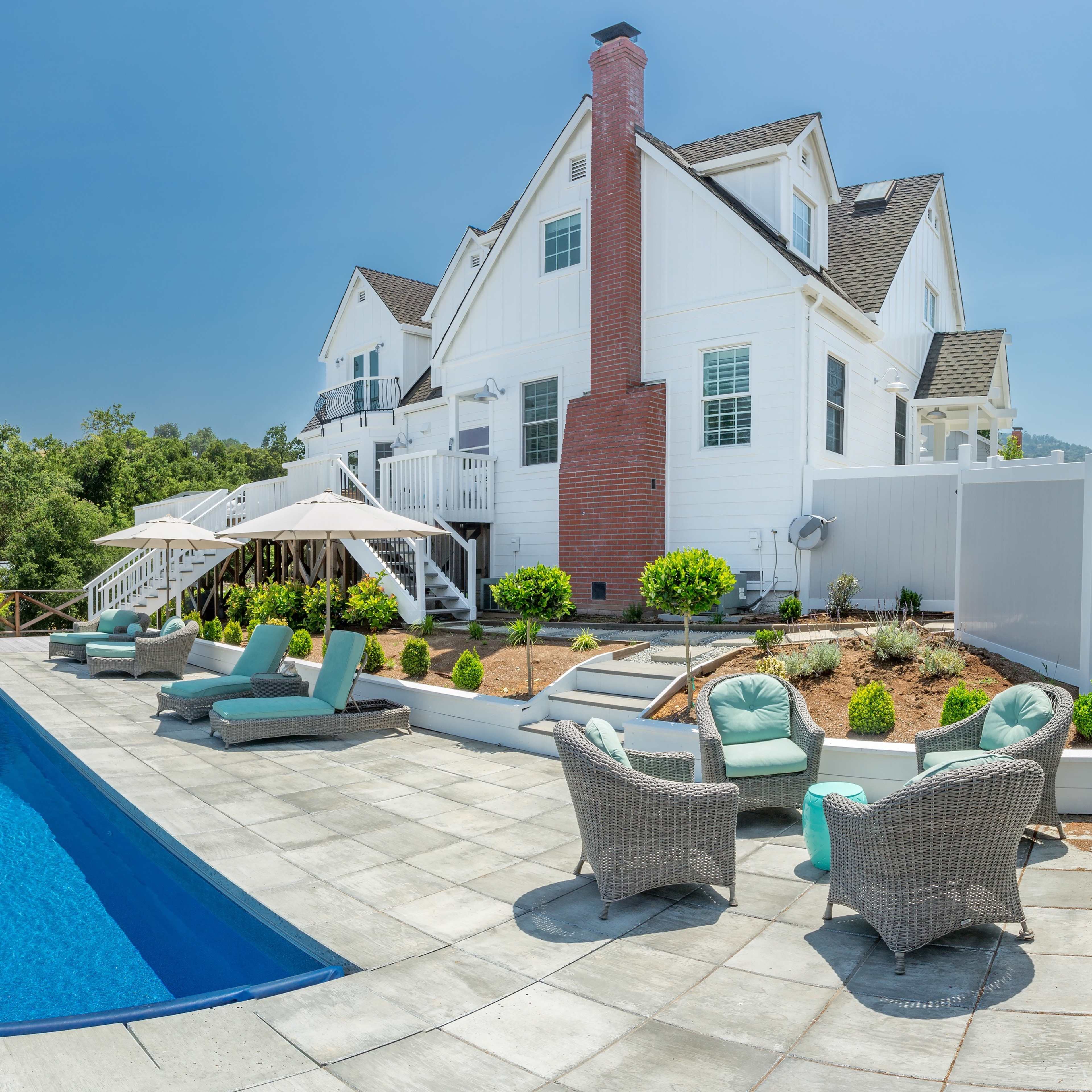 A well-maintained backyard features a rectangular swimming pool surrounded by lounge chairs and a neatly landscaped area with hedges and a two-story white house in the background.