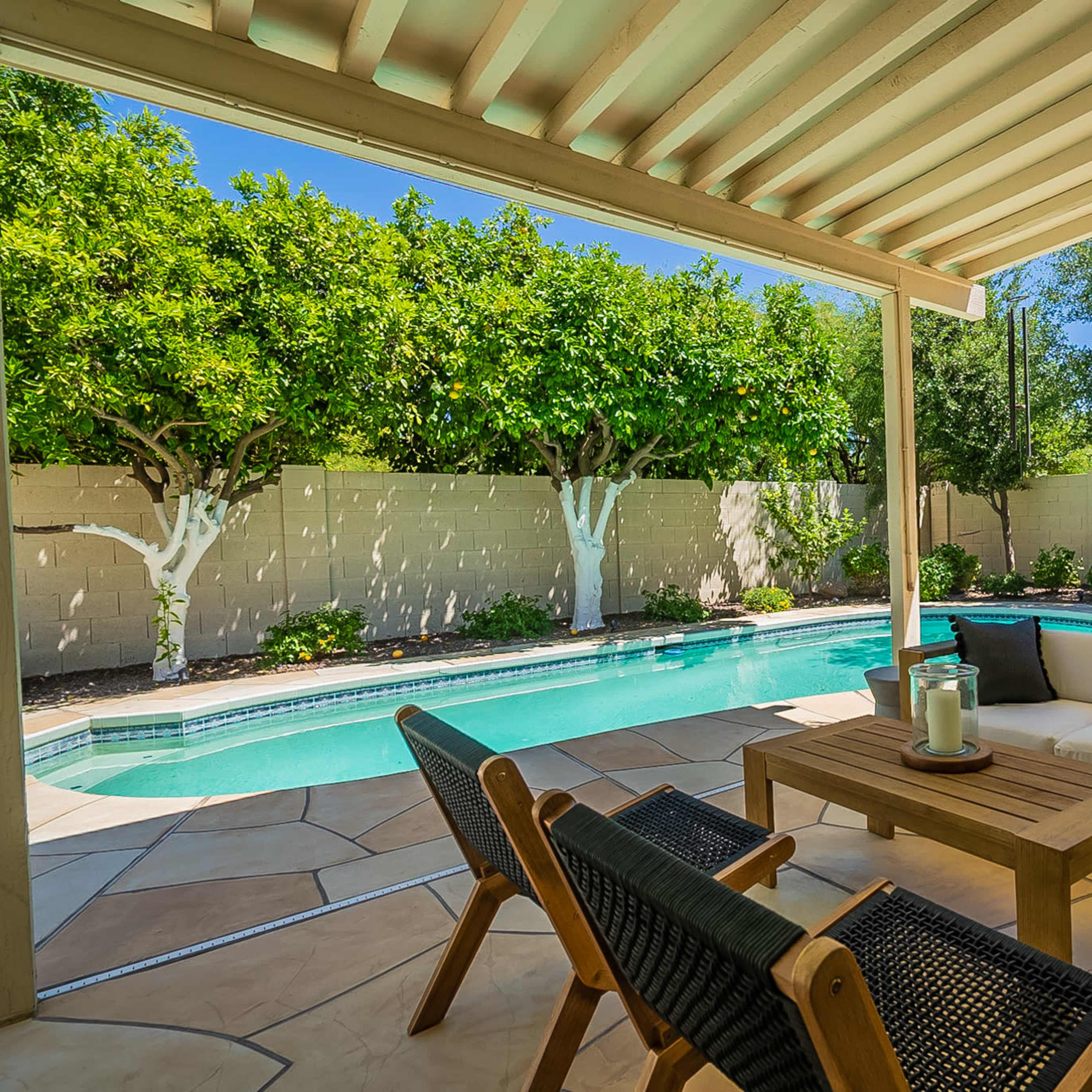 A patio area with a wooden seating arrangement overlooks a swimming pool surrounded by greenery and a stone fence.