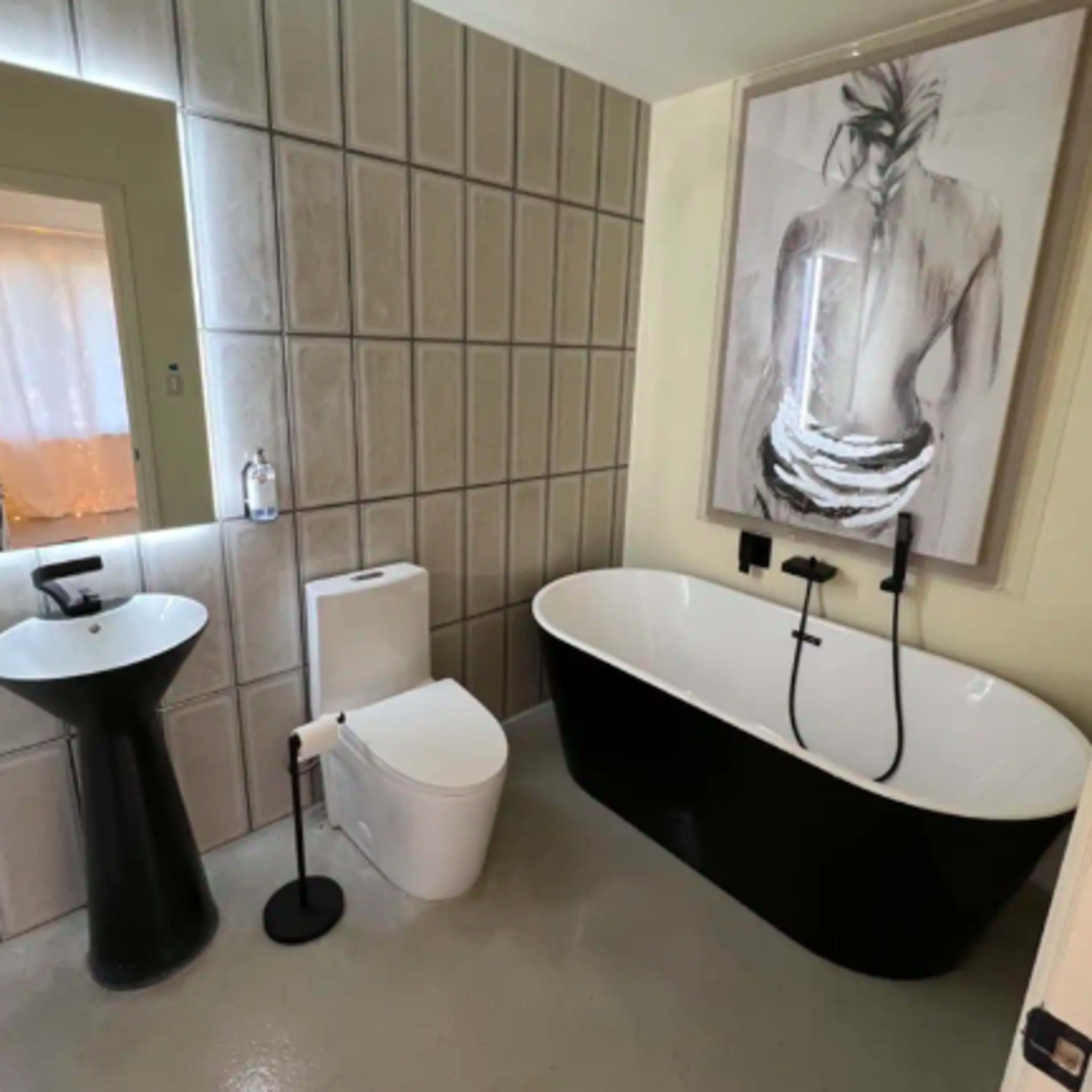 The bathroom features a freestanding black and white bathtub, a modern toilet, a pedestal sink, and a large framed artwork on the wall.
