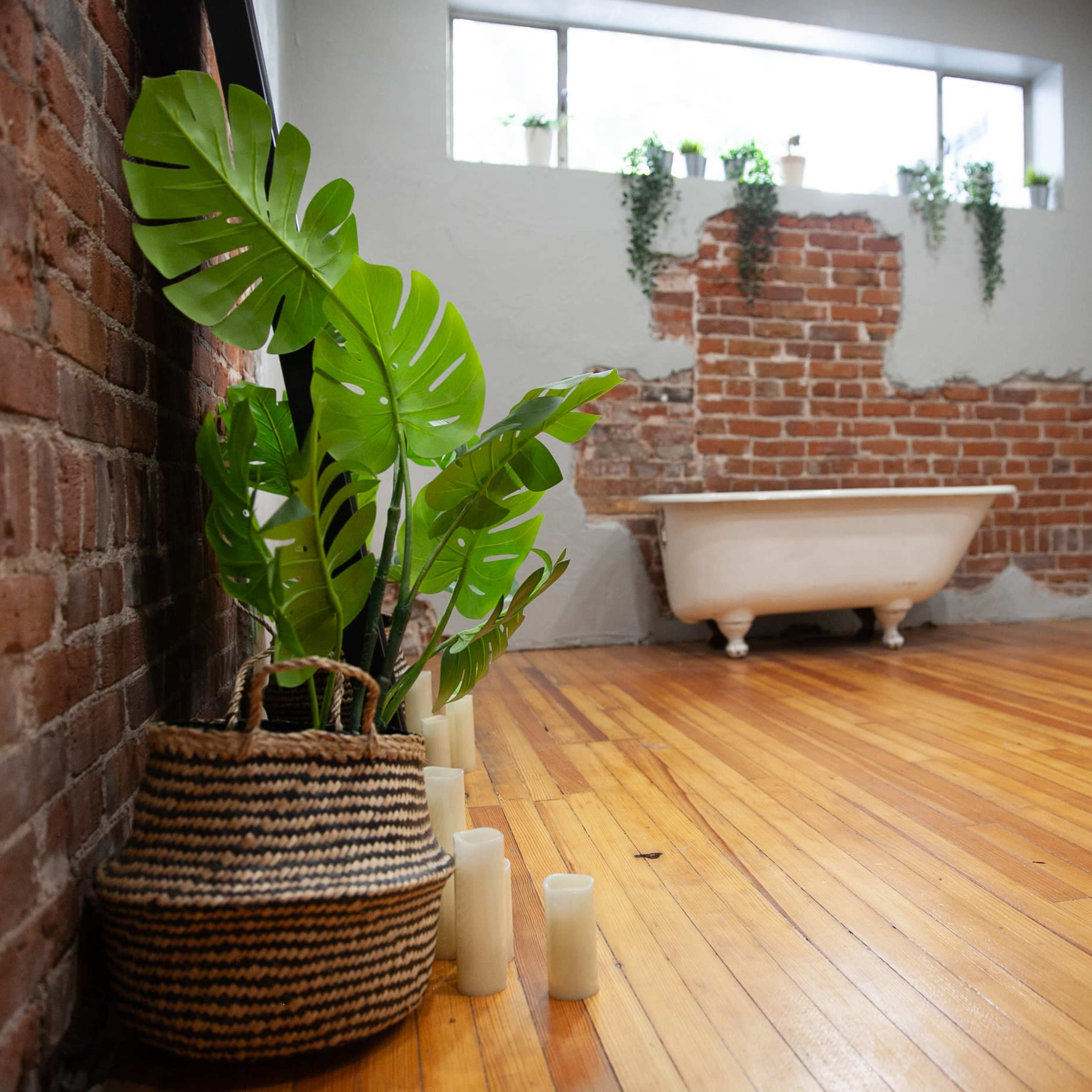 A room with exposed brick walls, a vintage bathtub, and potted plants beside a woven basket on polished wooden flooring.