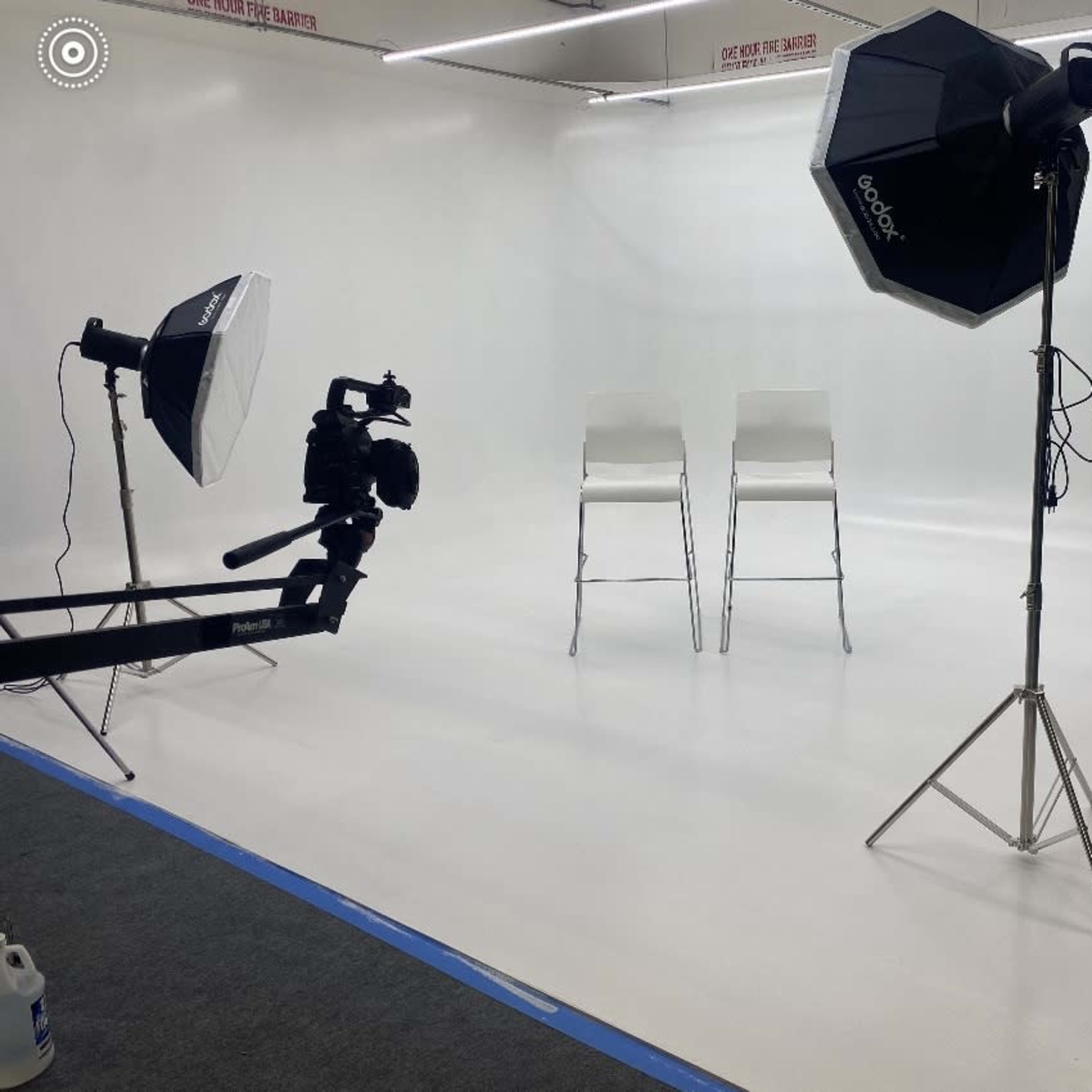 A photography studio with two empty chairs, lighting equipment, and a camera setup against a plain white backdrop.