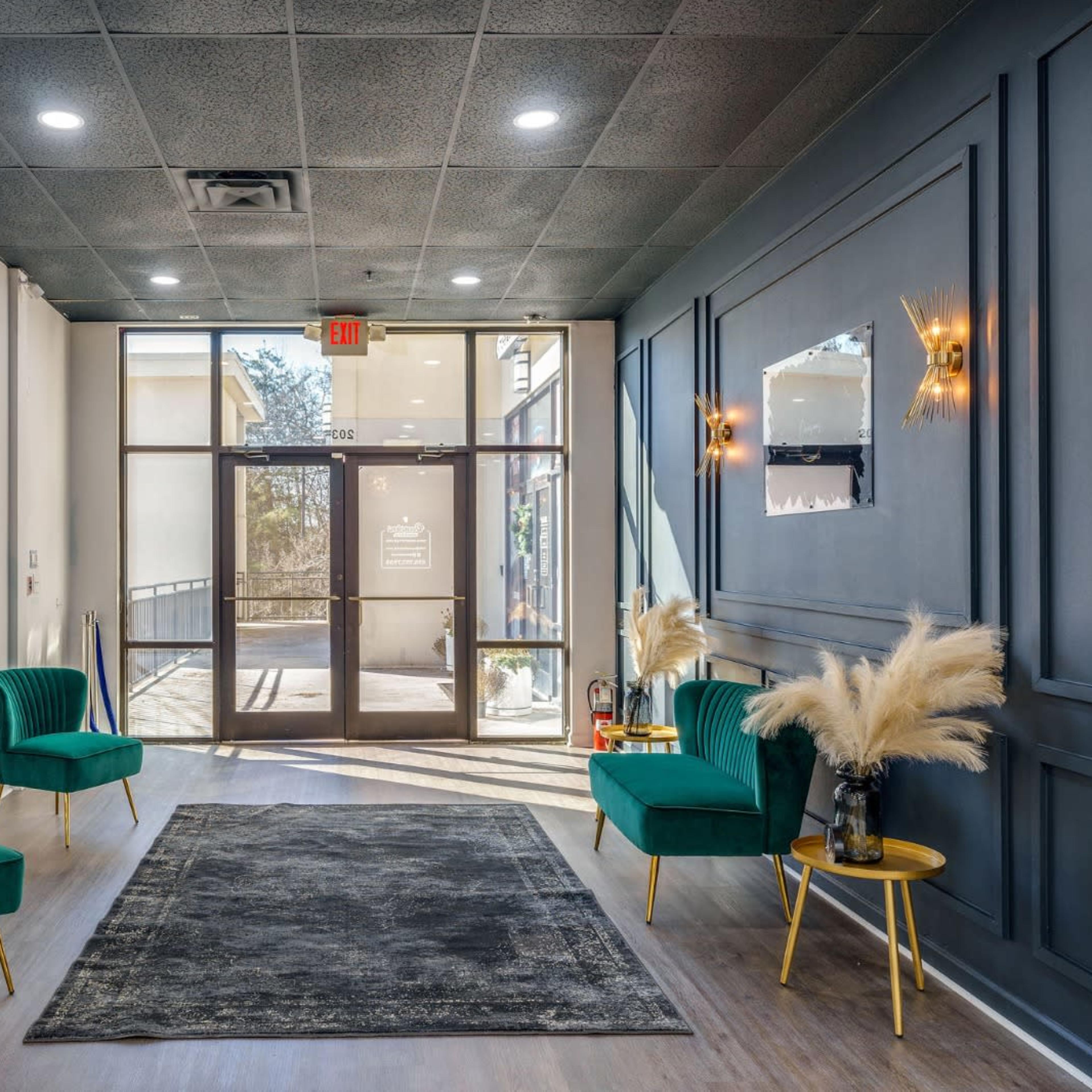 A modern lobby with two green chairs, a large mirror, and a decorative plant arrangement, illuminated by natural light from the entrance.