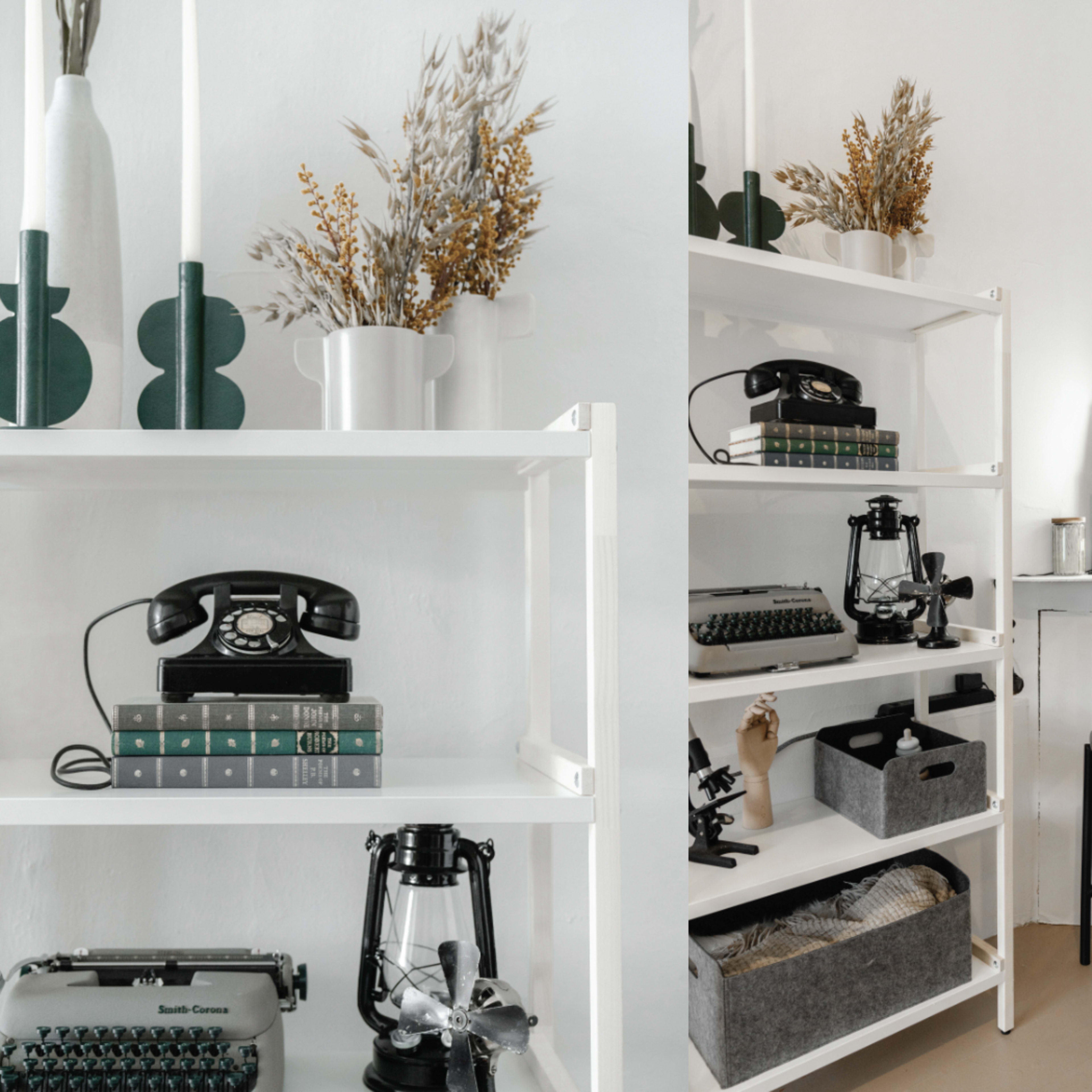 A neatly arranged interior space featuring a white shelf with vintage typewriters, decorative vases, and a retro telephone, alongside a cozy seating area with a framed artwork on the side.