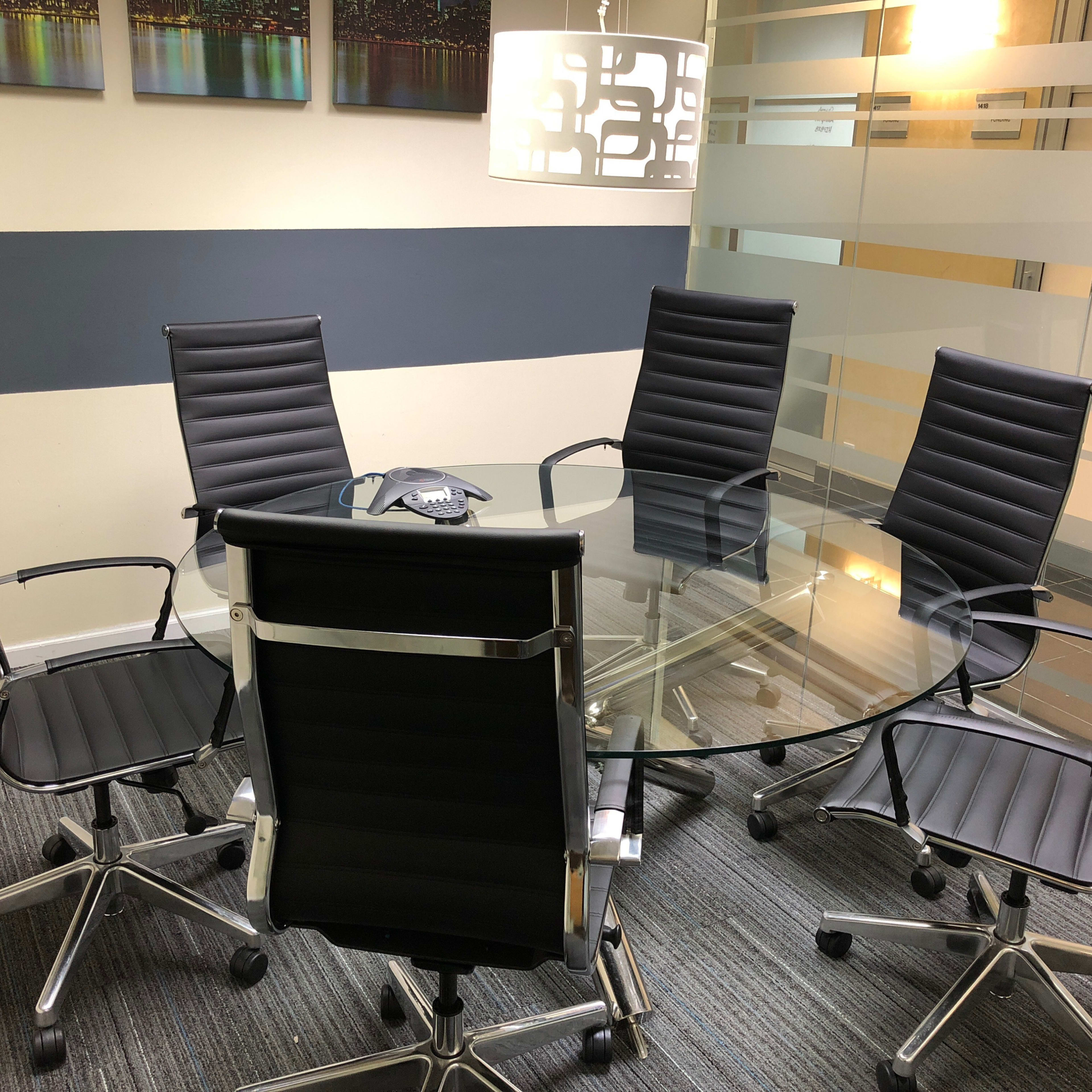 A glass conference table surrounded by five black office chairs is positioned in a well-lit meeting room with abstract artwork on the walls.