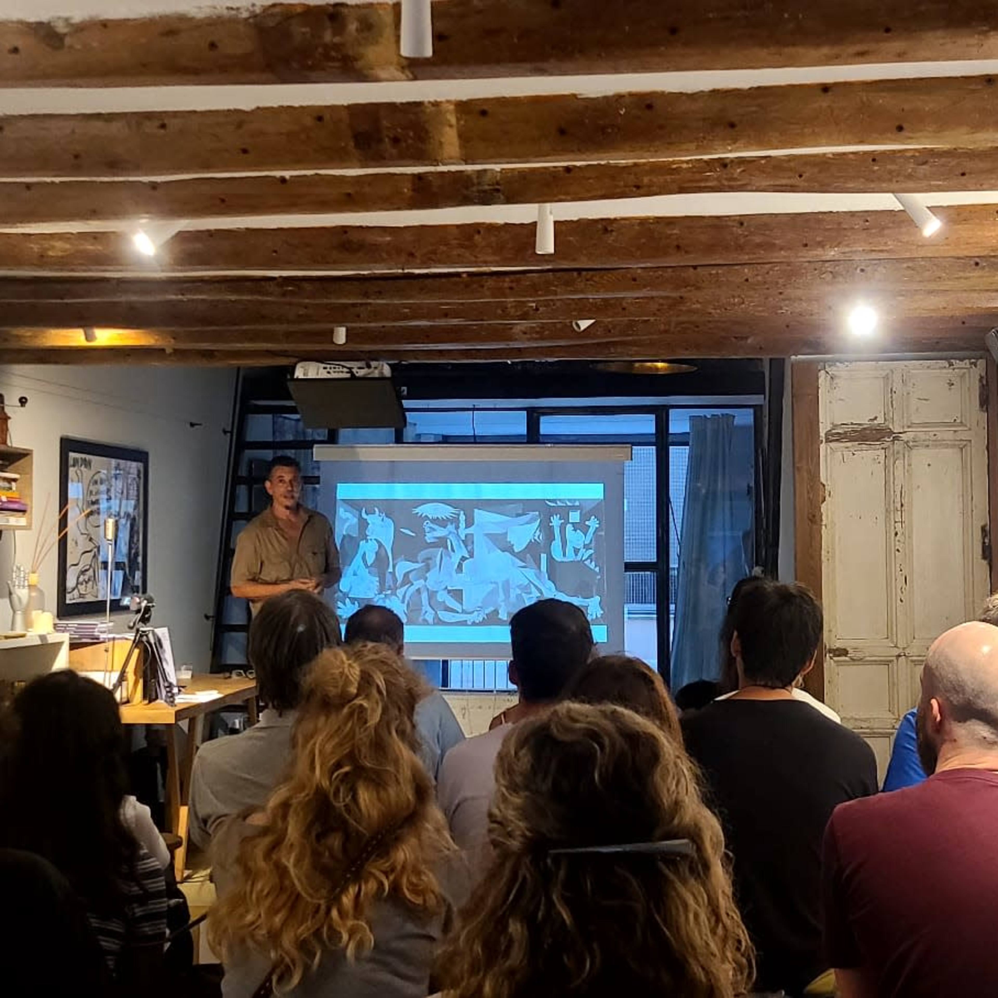 A presenter stands in front of an audience in a room with exposed wooden beams, delivering a talk while a projector displays images on a screen.