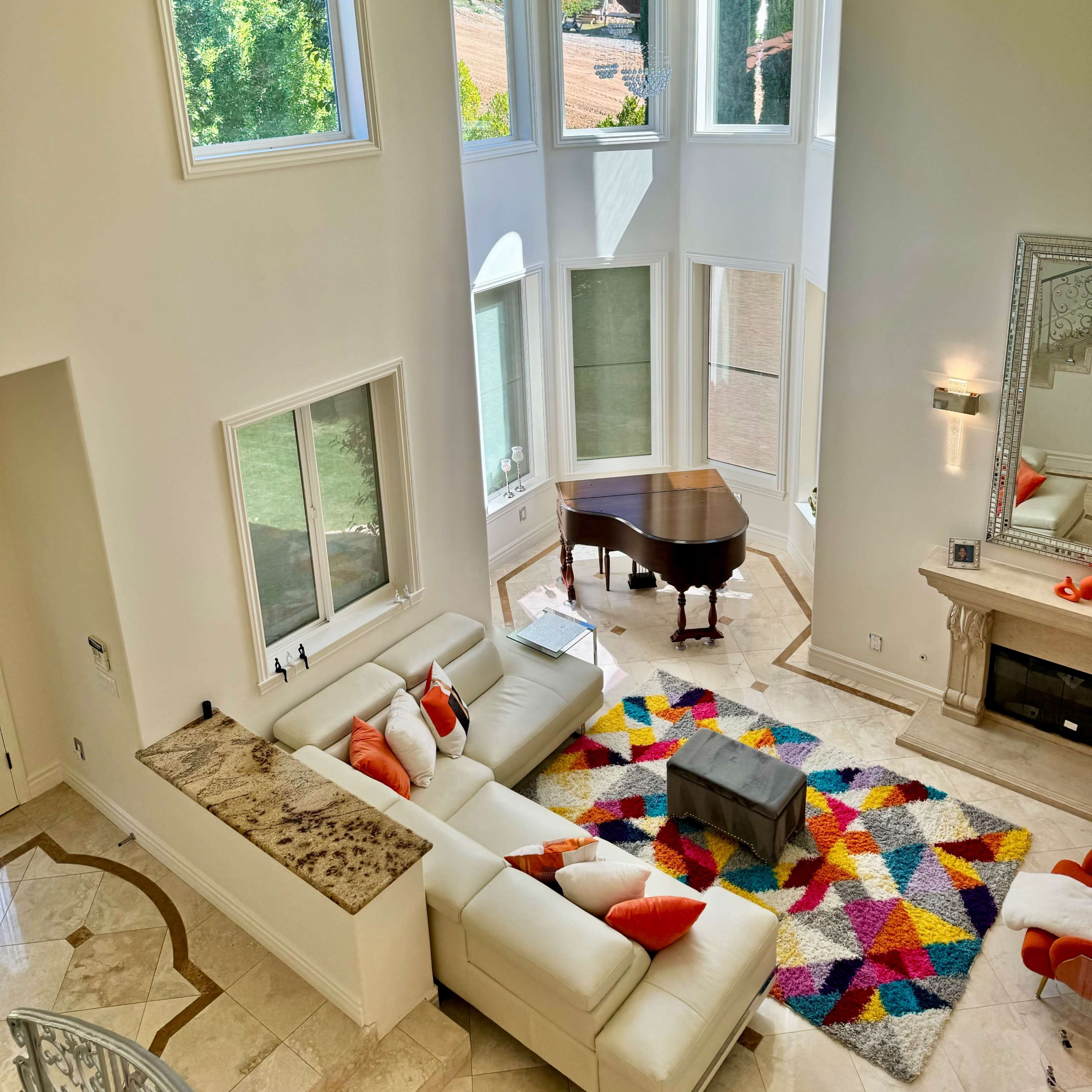 A spacious living room with a white sectional sofa, colorful rug, a grand piano, and large windows that let in natural light.