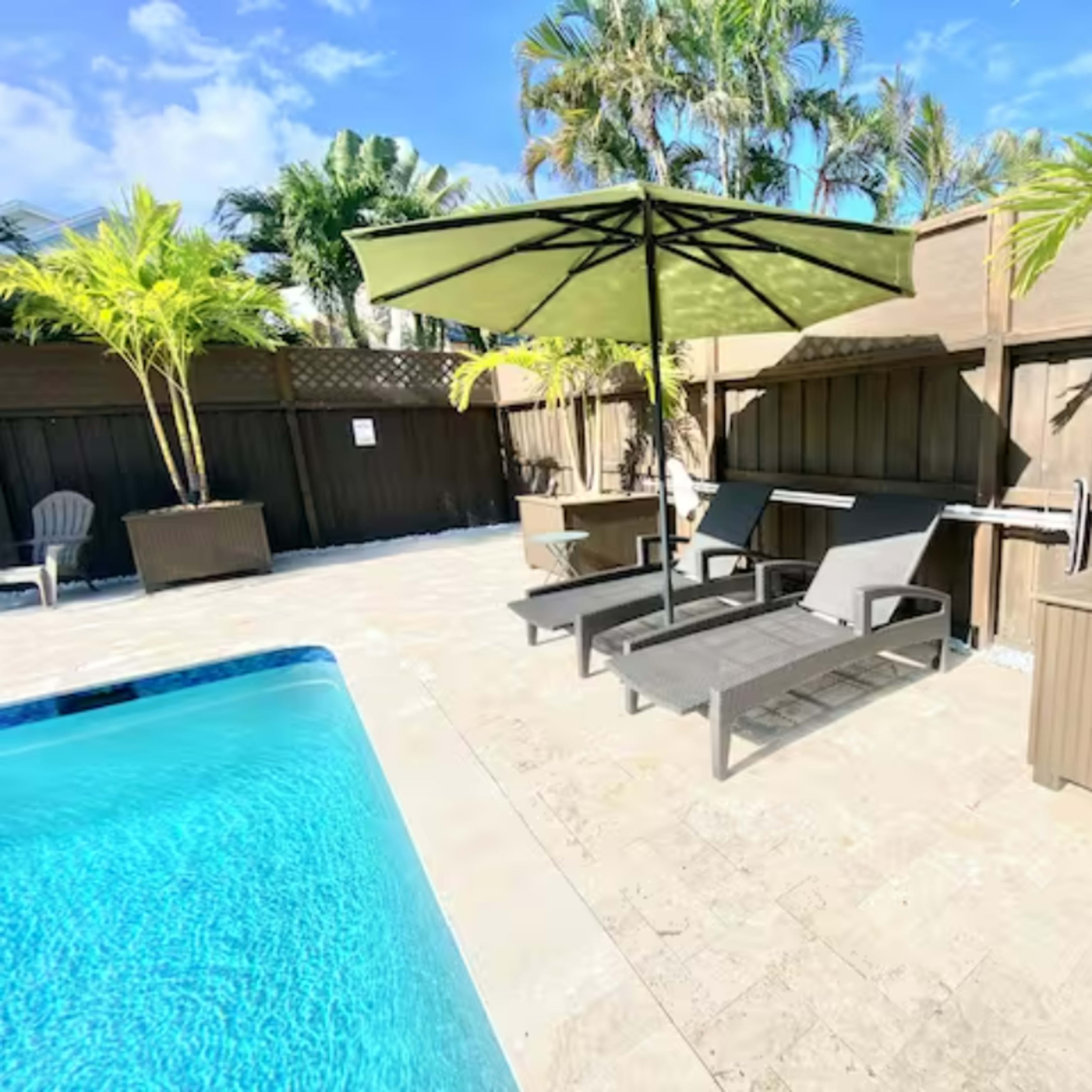The scene features a swimming pool area with two lounge chairs and an umbrella, surrounded by palm trees and a wooden fence.