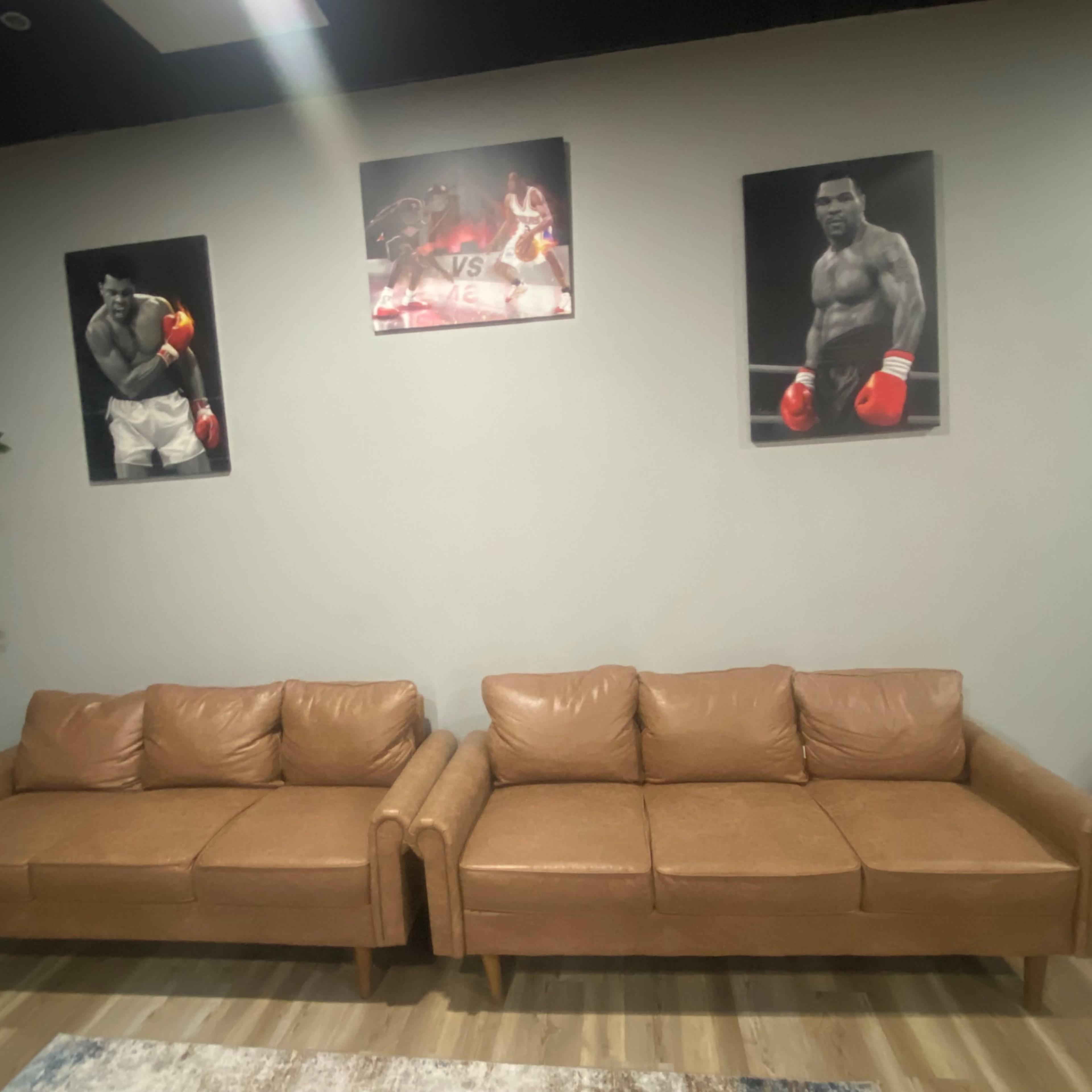 A pair of brown leather sofas is positioned against a gray wall that features two framed black-and-white boxing photographs.