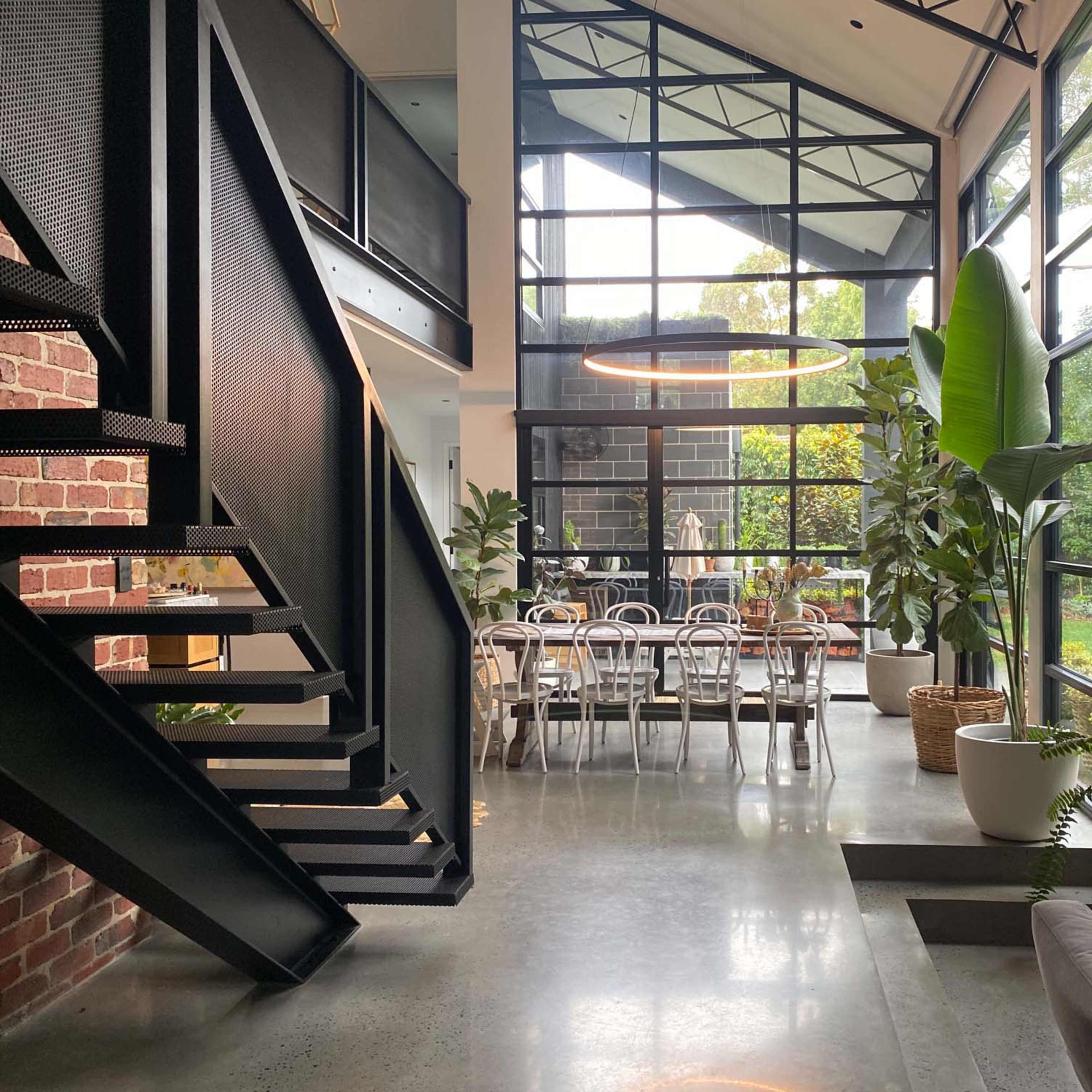 A modern open-concept living space with large windows, a staircase, and a dining area filled with plants.