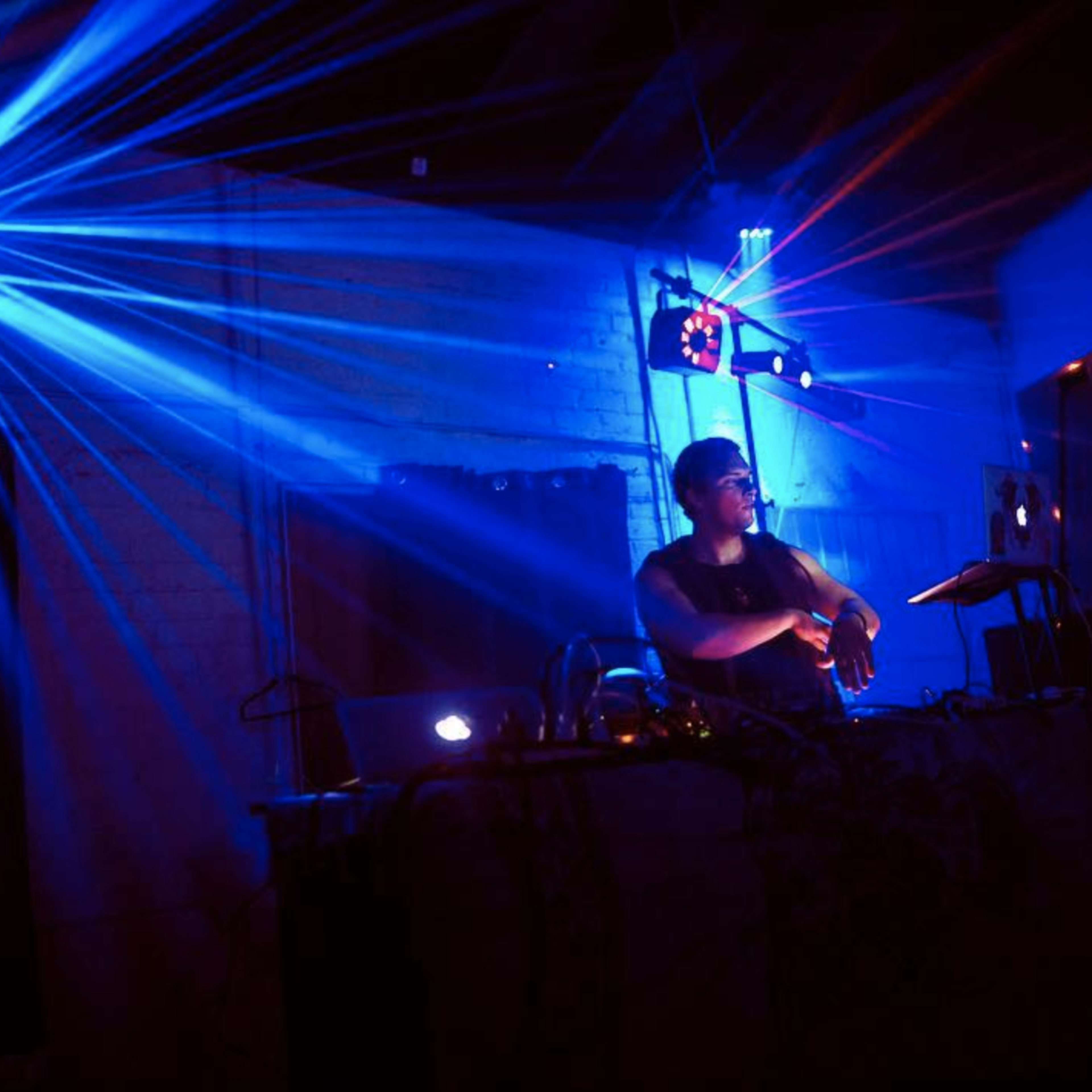 A DJ performs behind a mixing console with blue and purple laser lights illuminating the dark room.