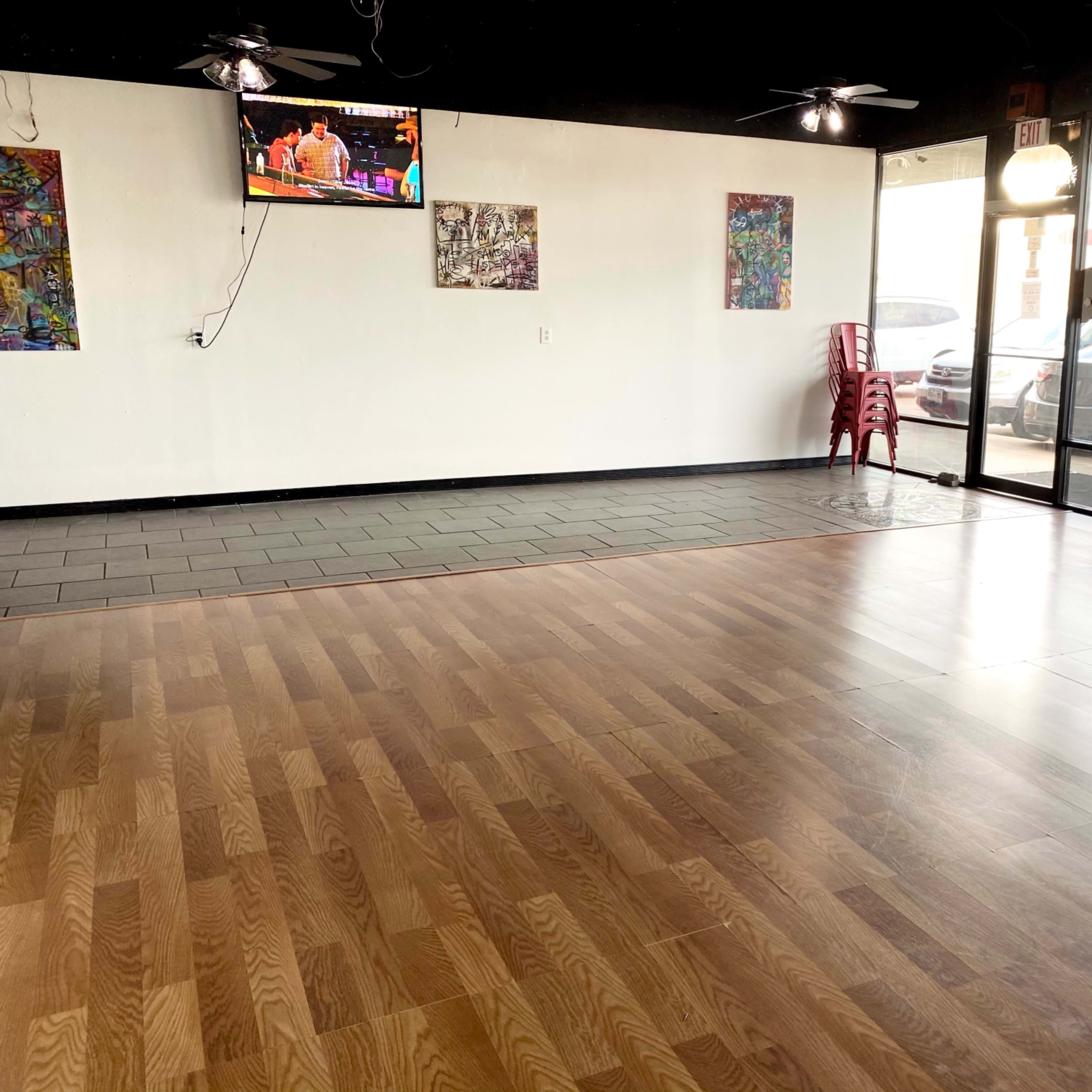 A spacious room with wood and tile flooring, a television mounted on the wall, and large windows displaying parked cars outside.