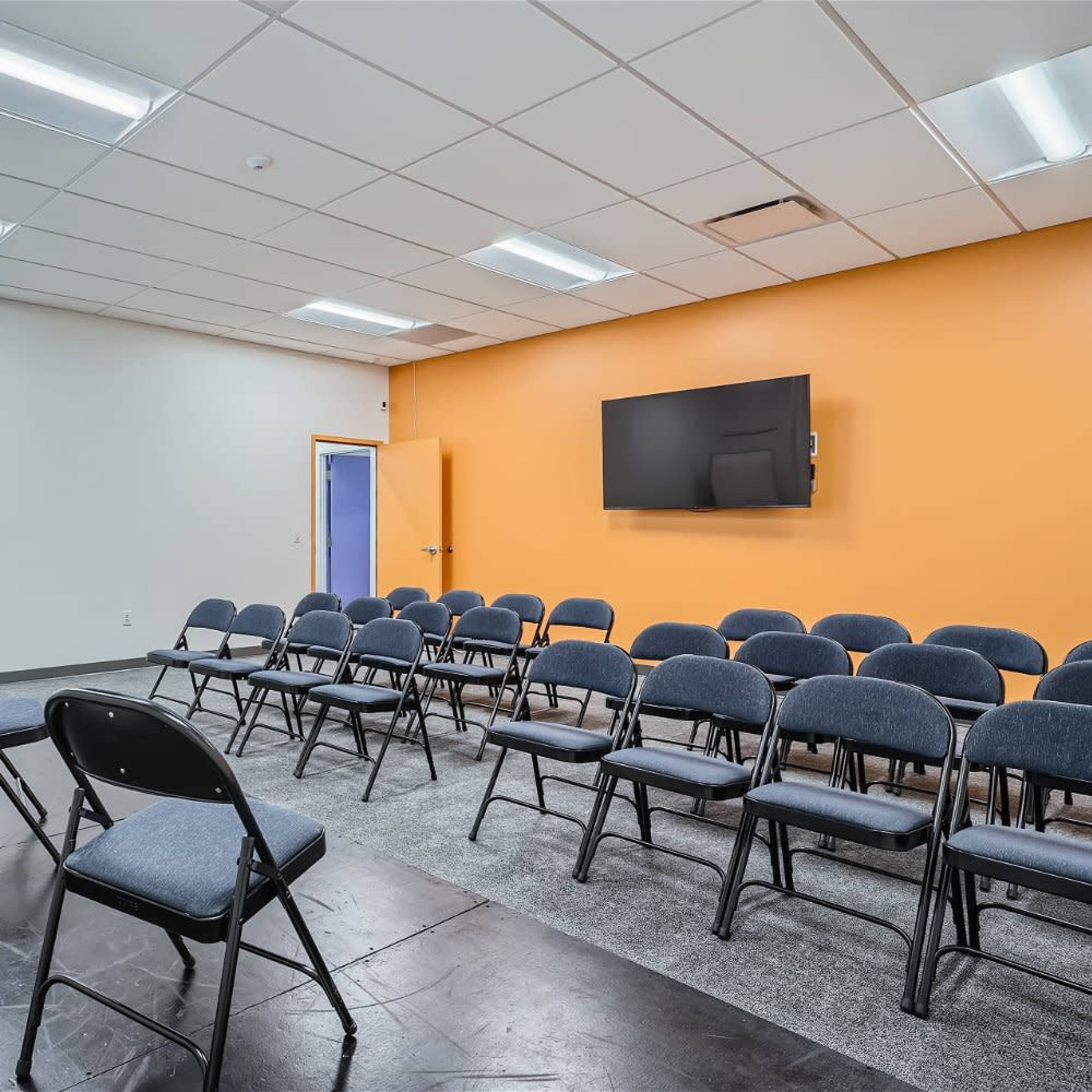A meeting room with rows of black chairs, a whiteboard, and a wall-mounted television, featuring an orange accent wall.