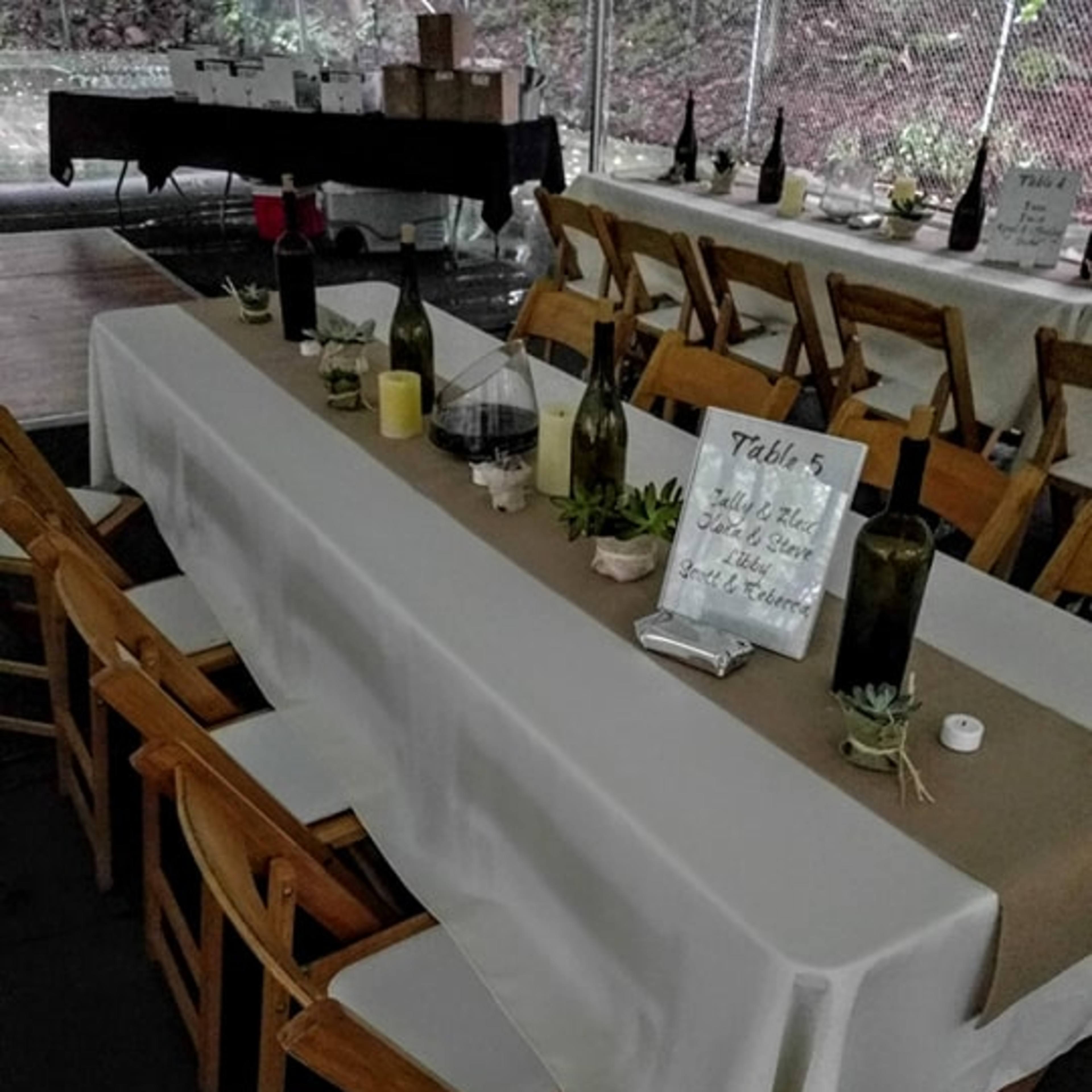 A long table is set for an event, featuring bottles, candles, and a sign indicating "Table 5," in a partially enclosed space.