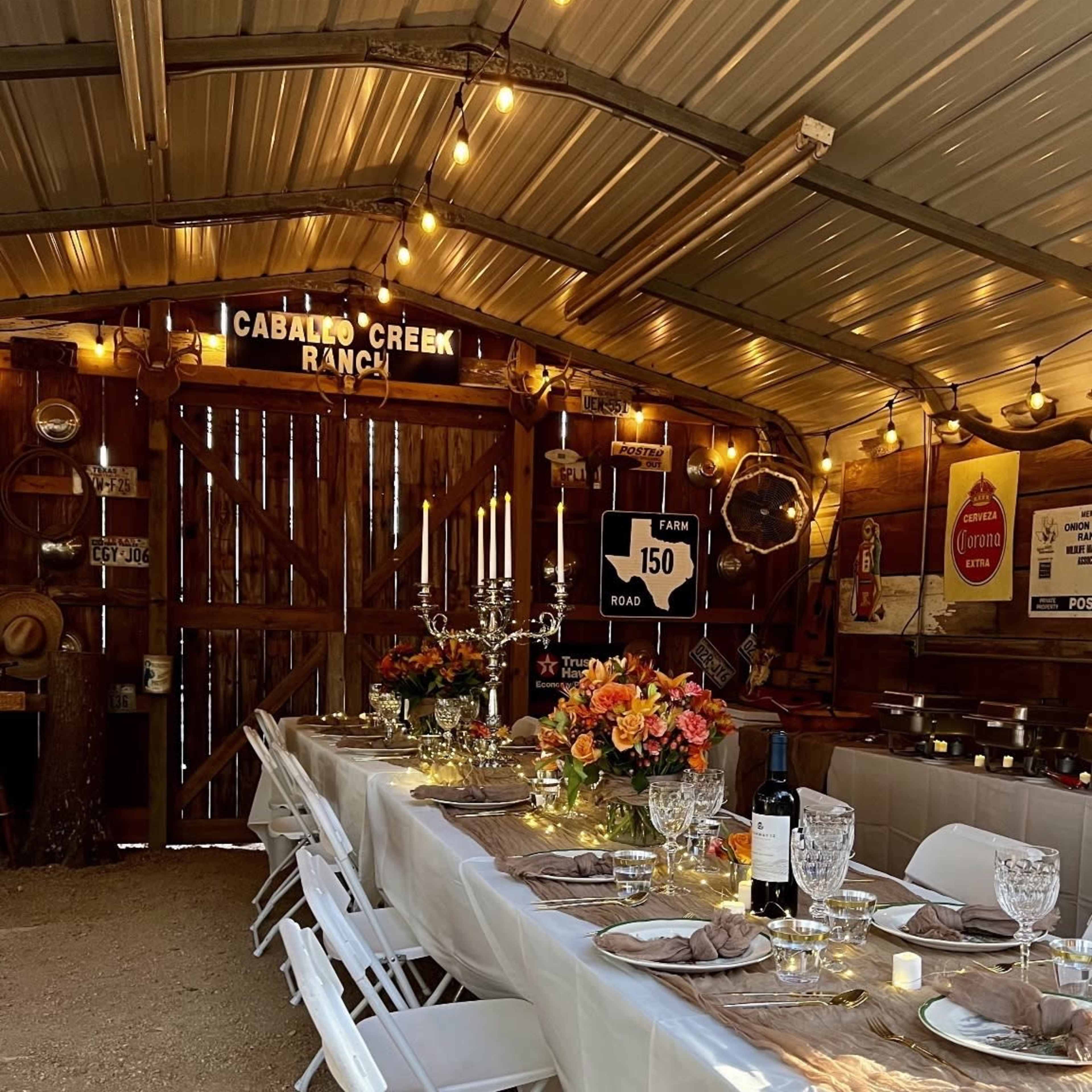 A long dining table is set with floral centerpieces and elegant tableware inside a rustic barn, adorned with string lights and vintage signs.