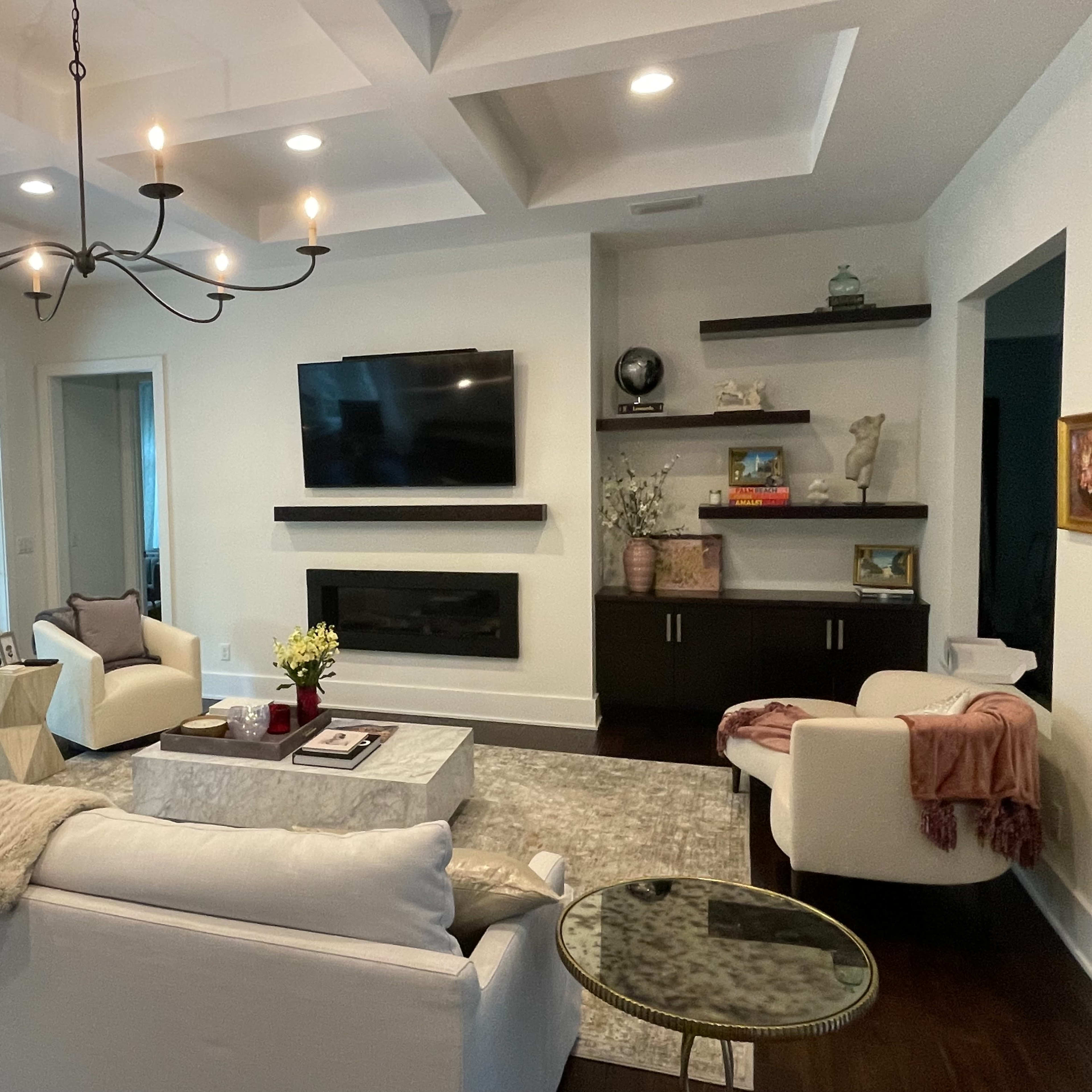 A modern living room with a white sofa, a coffee table, a television mounted on the wall, and a chandelier, all arranged around a space that features large windows and a view of greenery outside.