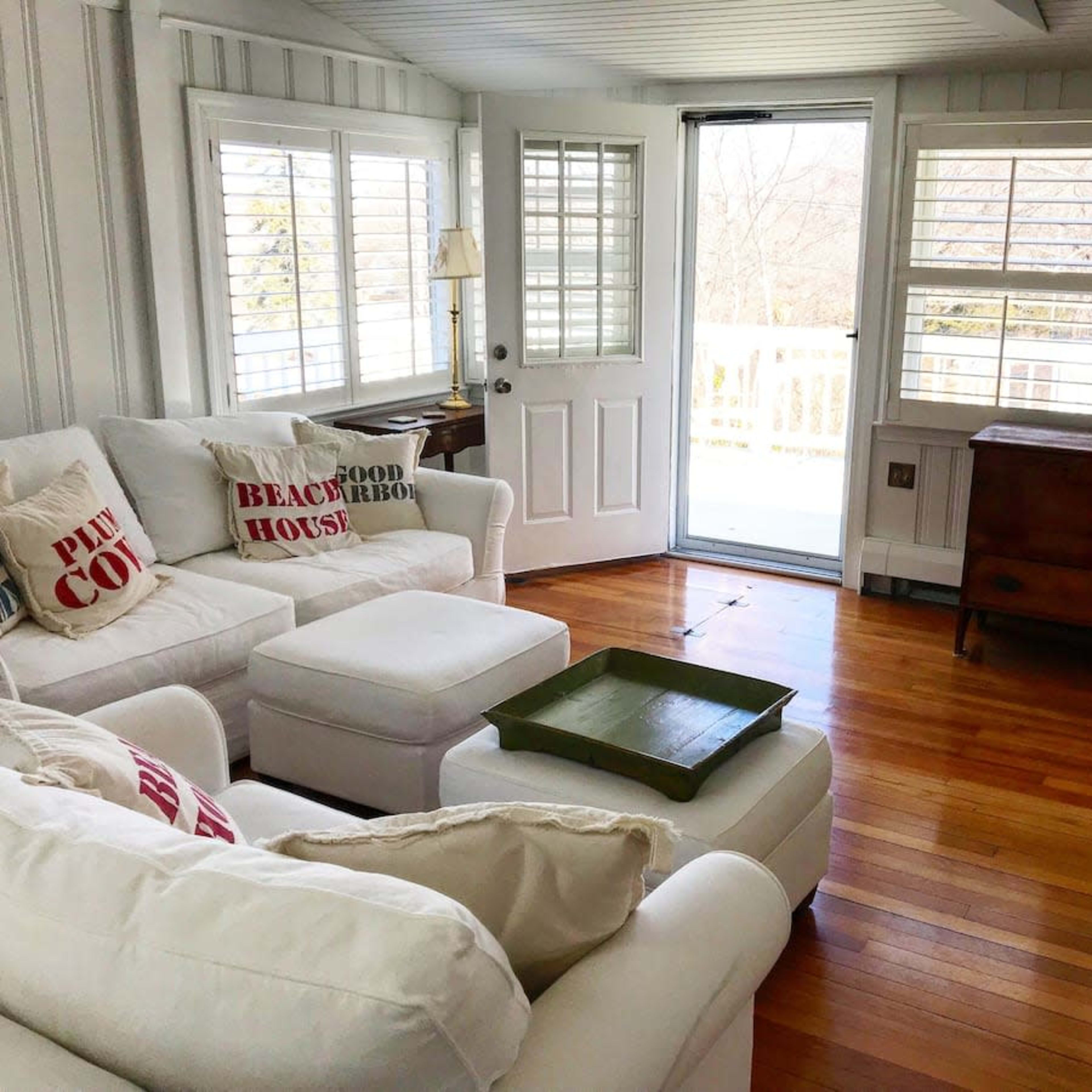 A bright, airy living room with a white sofa, decorative cushions, a wooden chest, and large windows that open to a balcony.