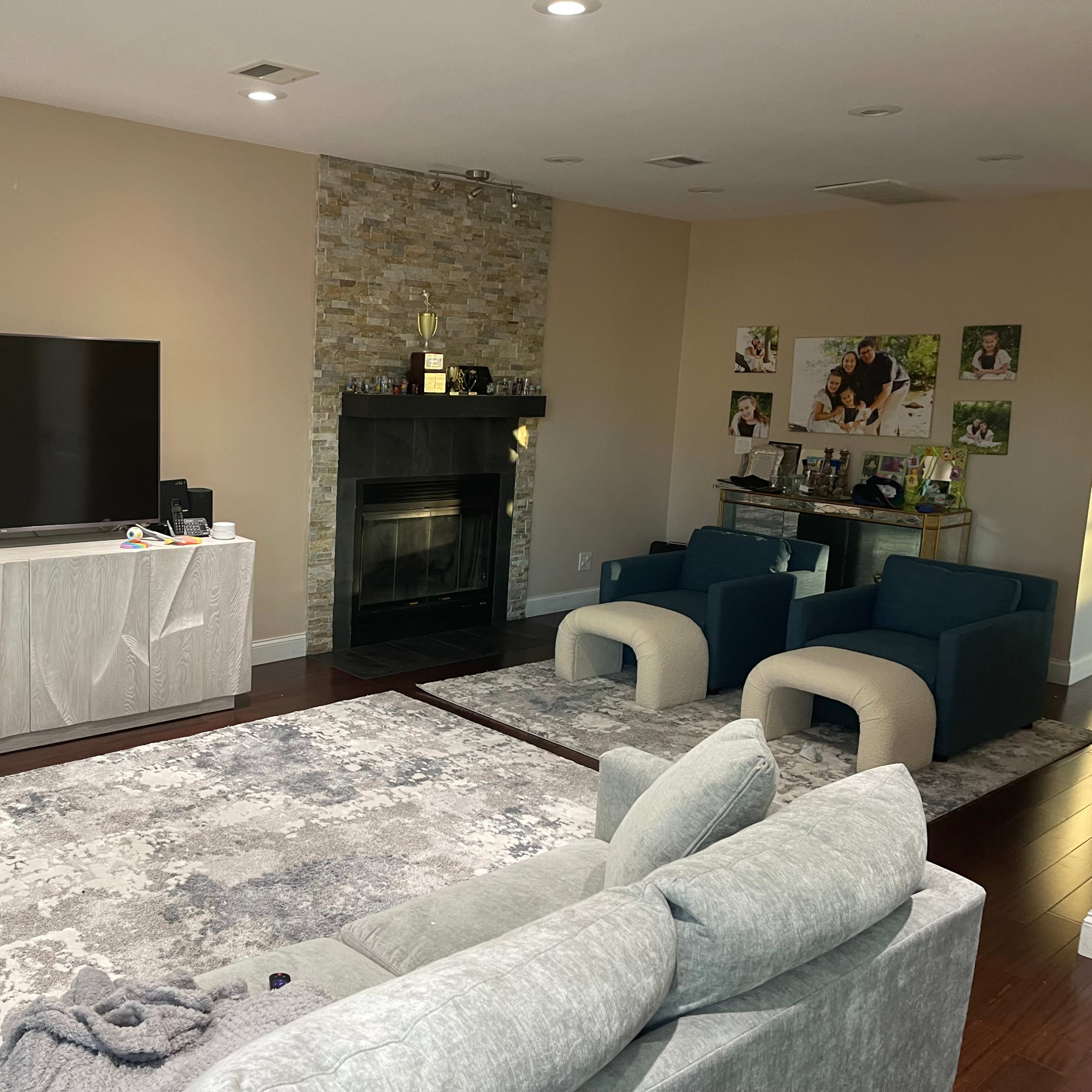 A modern living room features a large TV mounted on the wall, a stone fireplace, and two blue armchairs facing a light-colored sofa on a textured area rug.