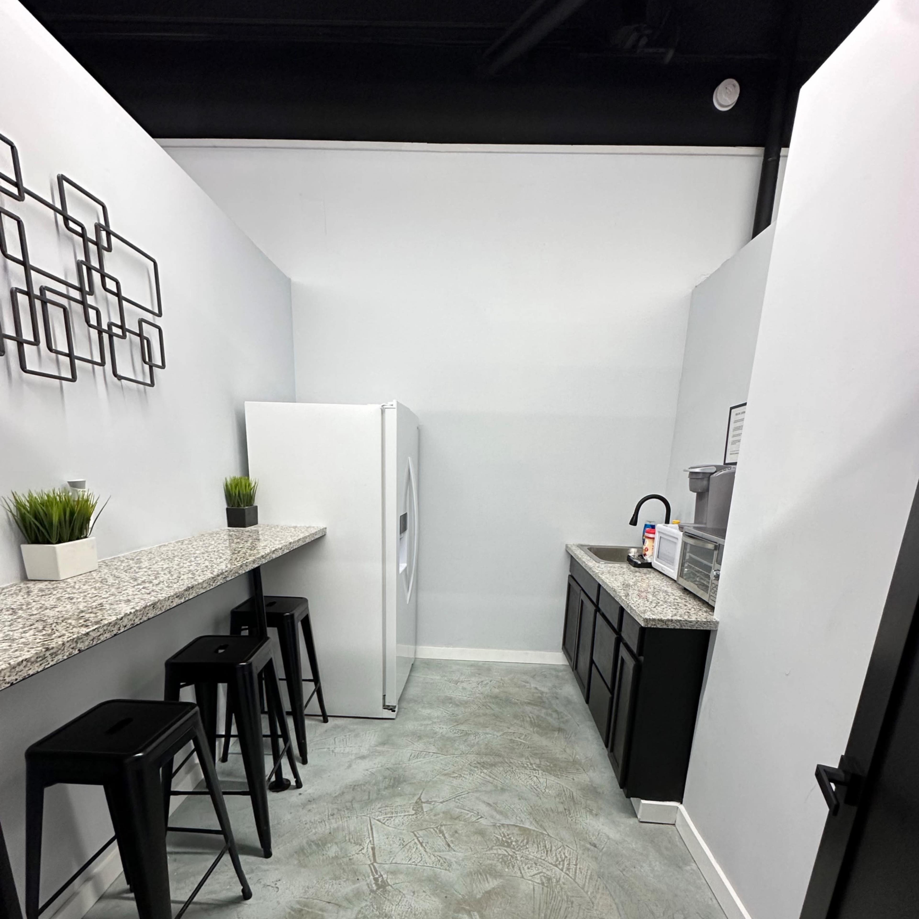 A minimalist kitchen area featuring a white refrigerator, dark cabinets, a countertop with stools, and a simple wall decor.