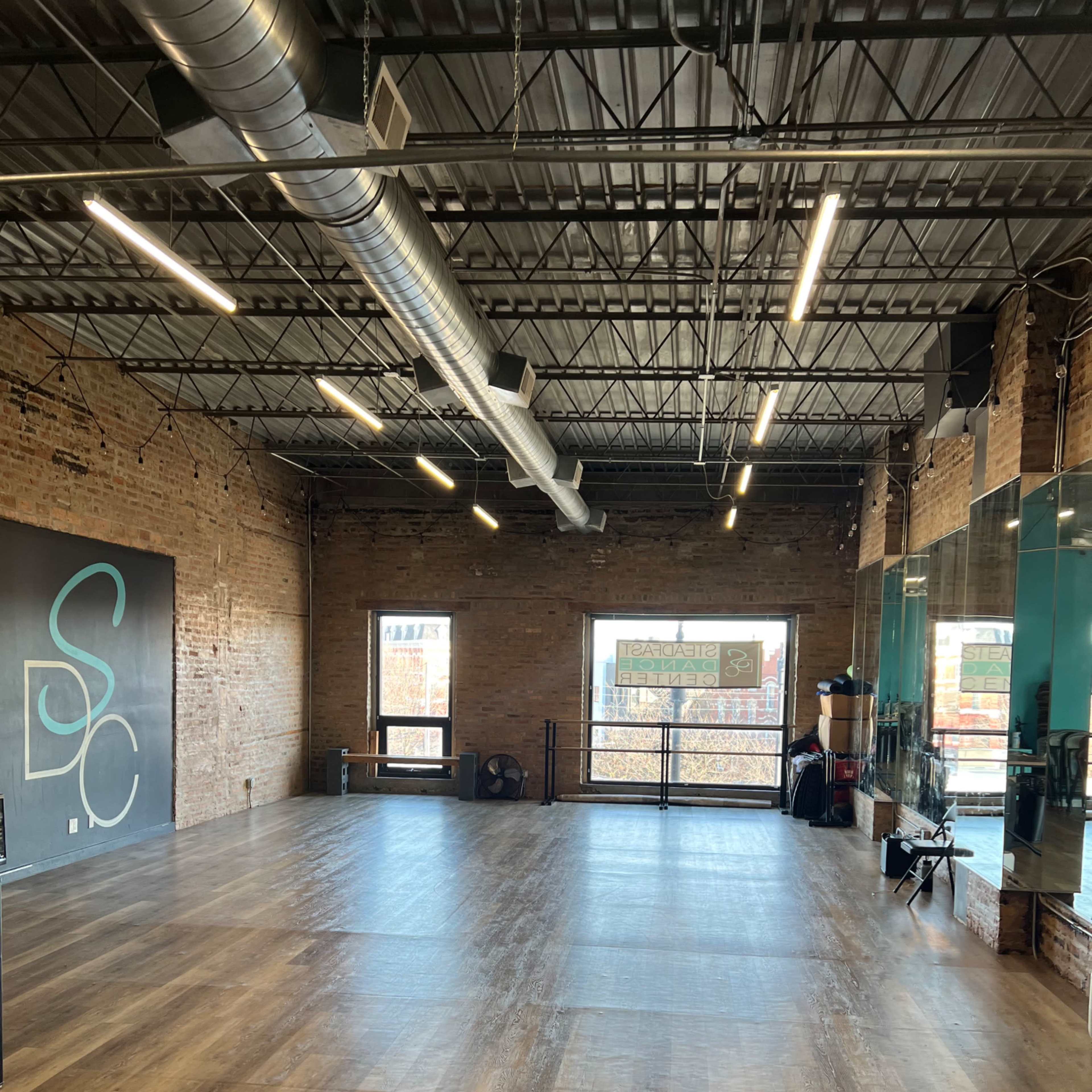 A spacious studio with wooden flooring, exposed brick walls, large windows, and mirrors on one side.