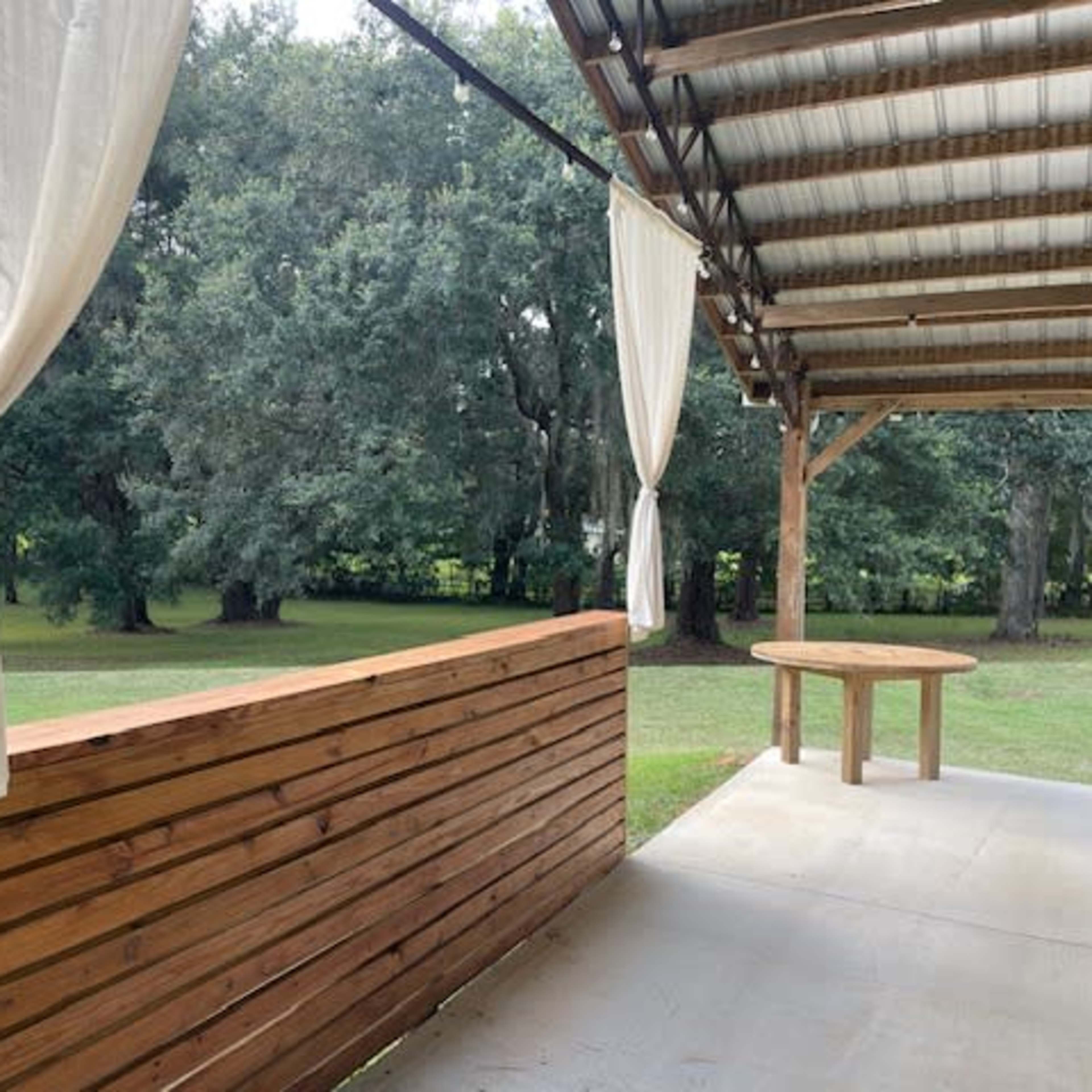 A wooden structure with a horizontal slatted design and a round table is set under a covered porch, leading to a grassy area with trees in the background.
