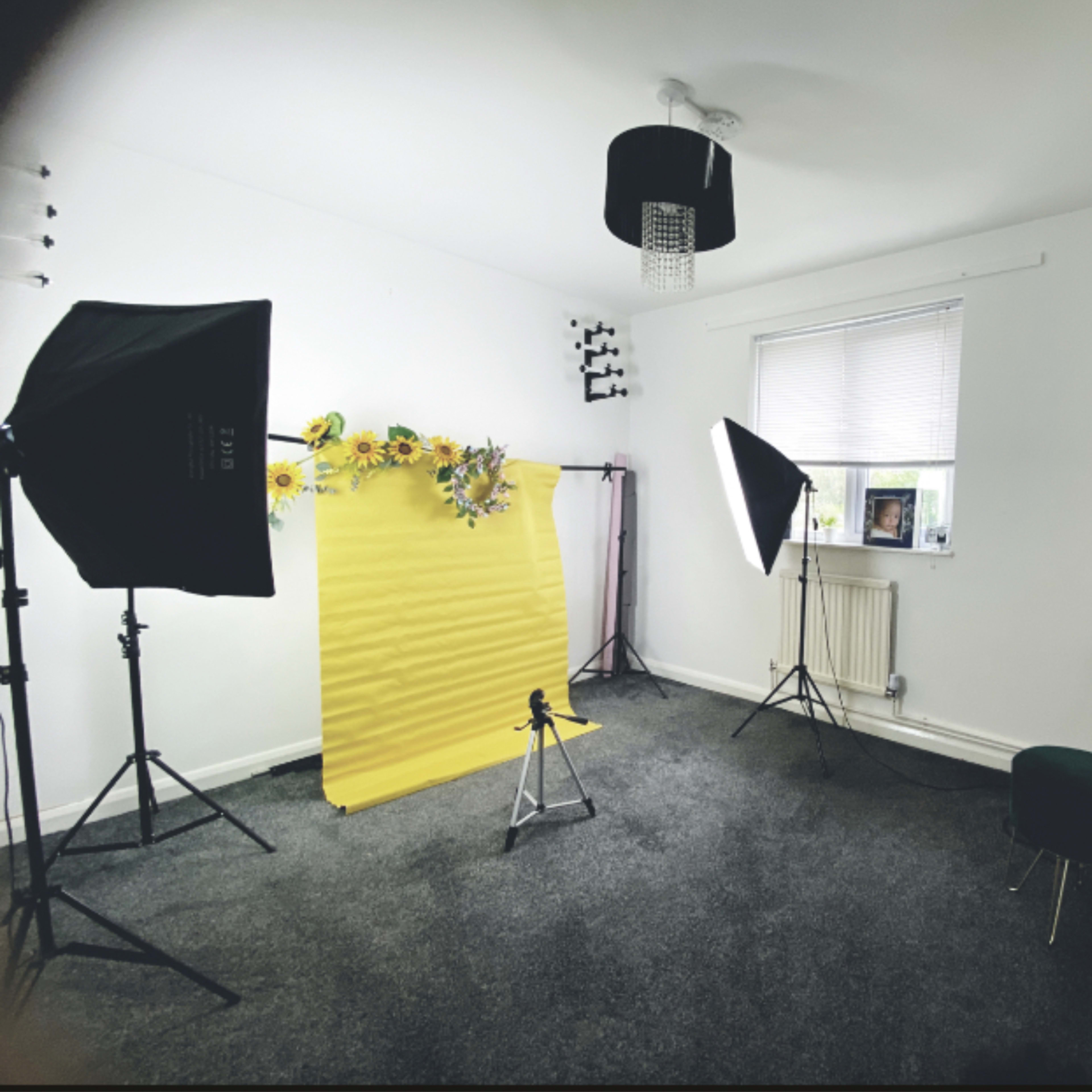 A photo studio setup with a yellow backdrop, two softbox lights, and a small green chair in a room with white walls and a carpeted floor.