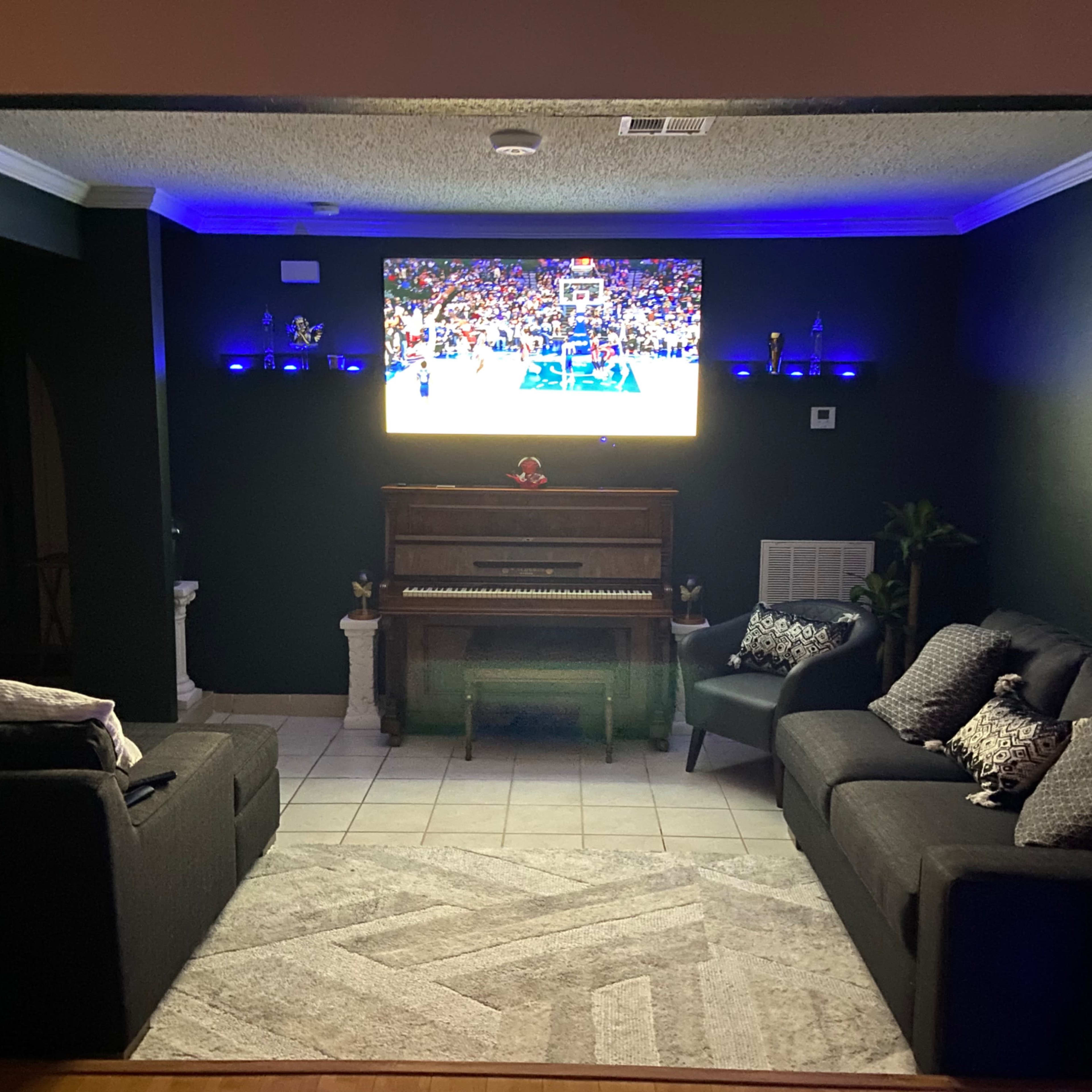 A living room features two sofas, a piano, and a television displaying a basketball game, with ambient lighting along the walls.