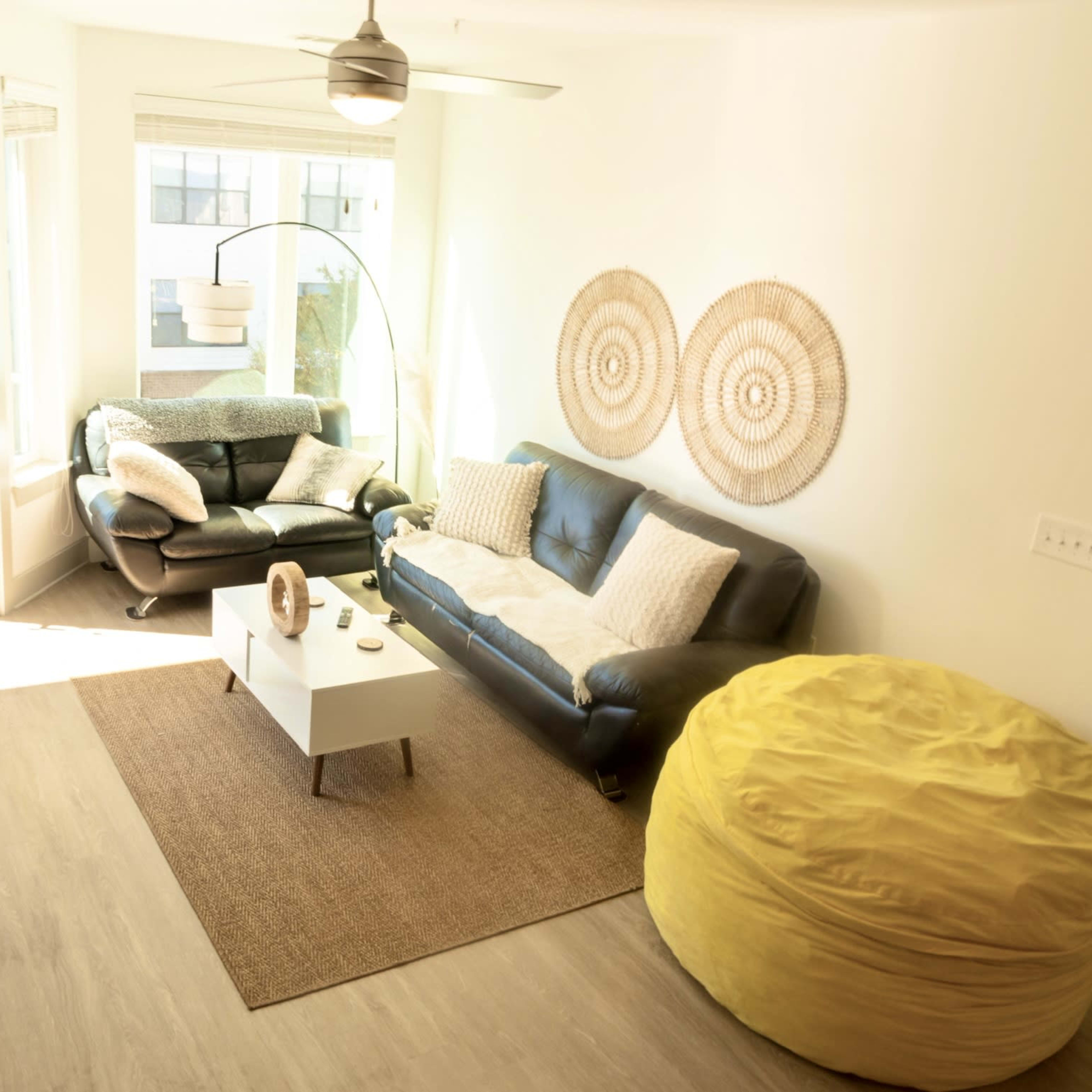 A bright living room with a leather sofa, a round coffee table, a large yellow bean bag, and decorative wall art made of woven materials.