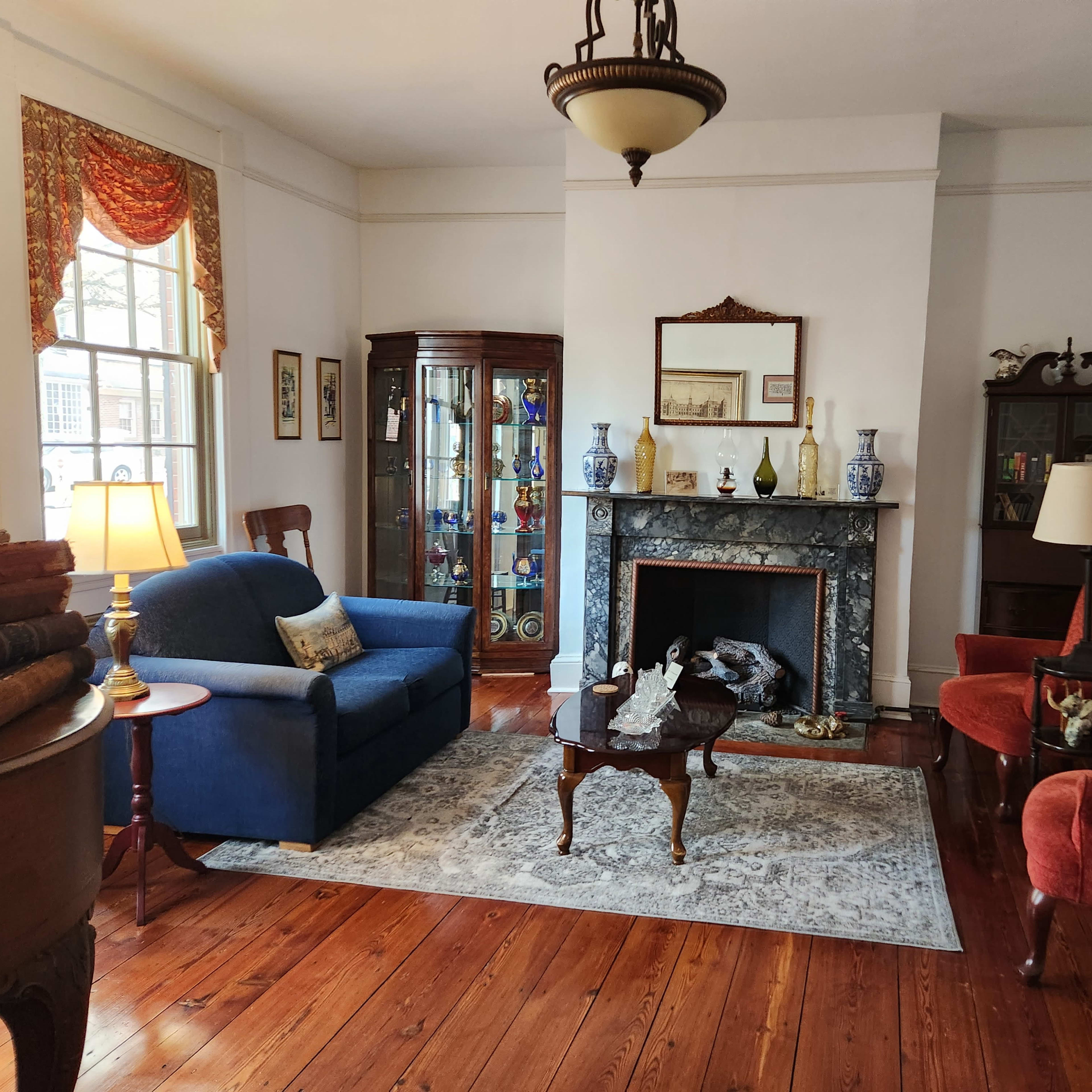 A living room with a blue couch, two red armchairs, a wooden coffee table on a patterned rug, and a fireplace with decorative items.