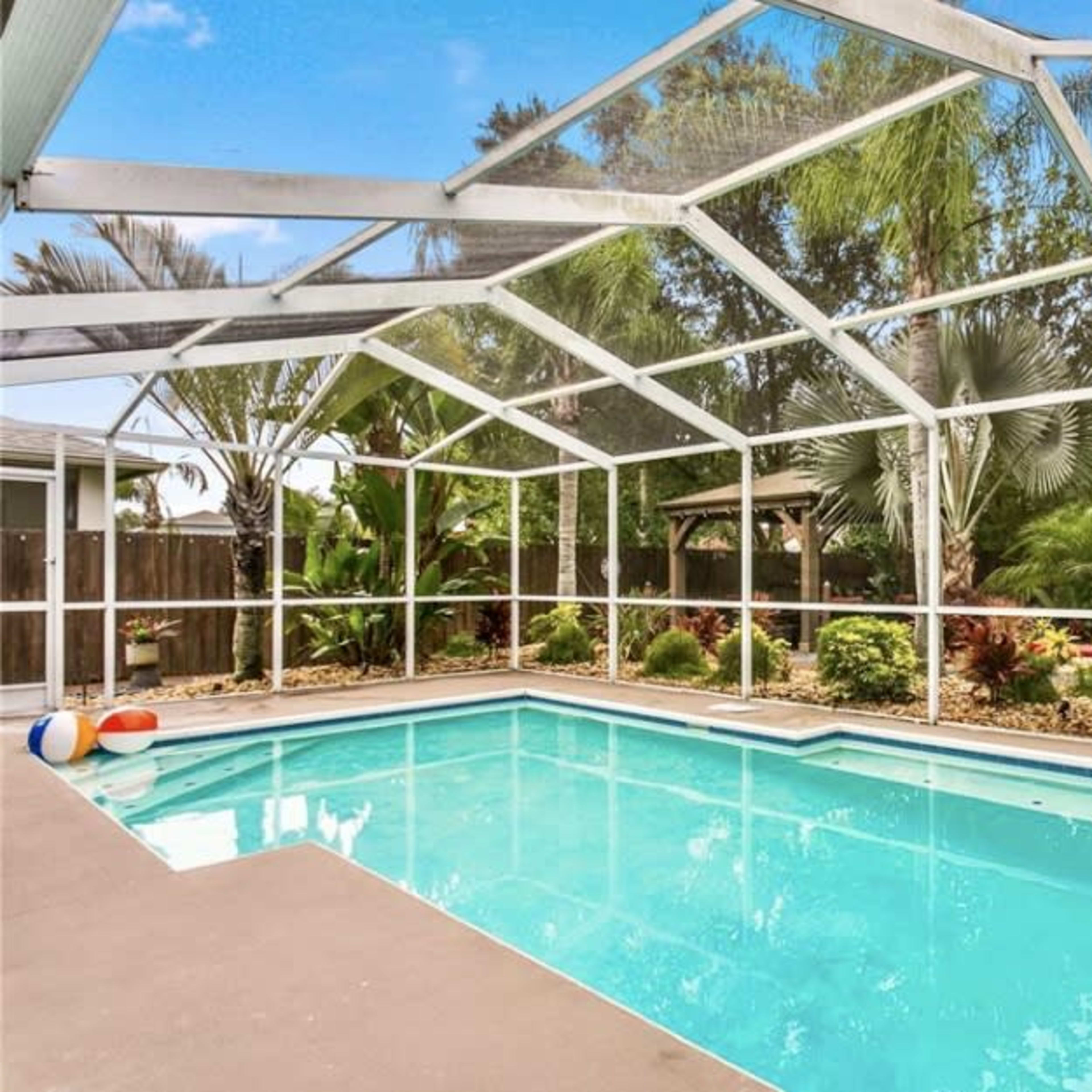 A screened-in swimming pool is surrounded by tropical plants and landscaping in a residential backyard.