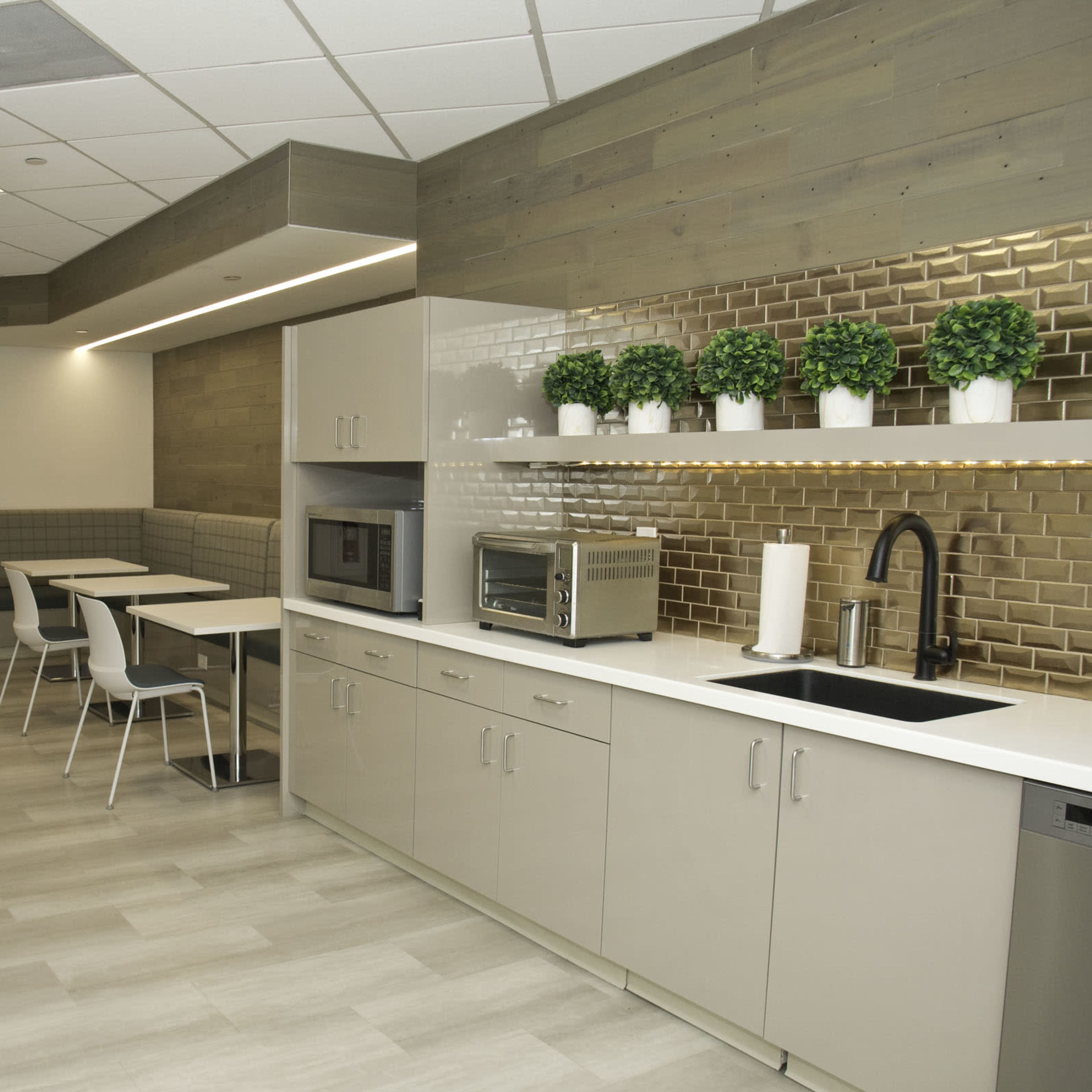 A modern kitchen area featuring a countertop, microwave, sink, and several tables and chairs against a wall with decorative plants.