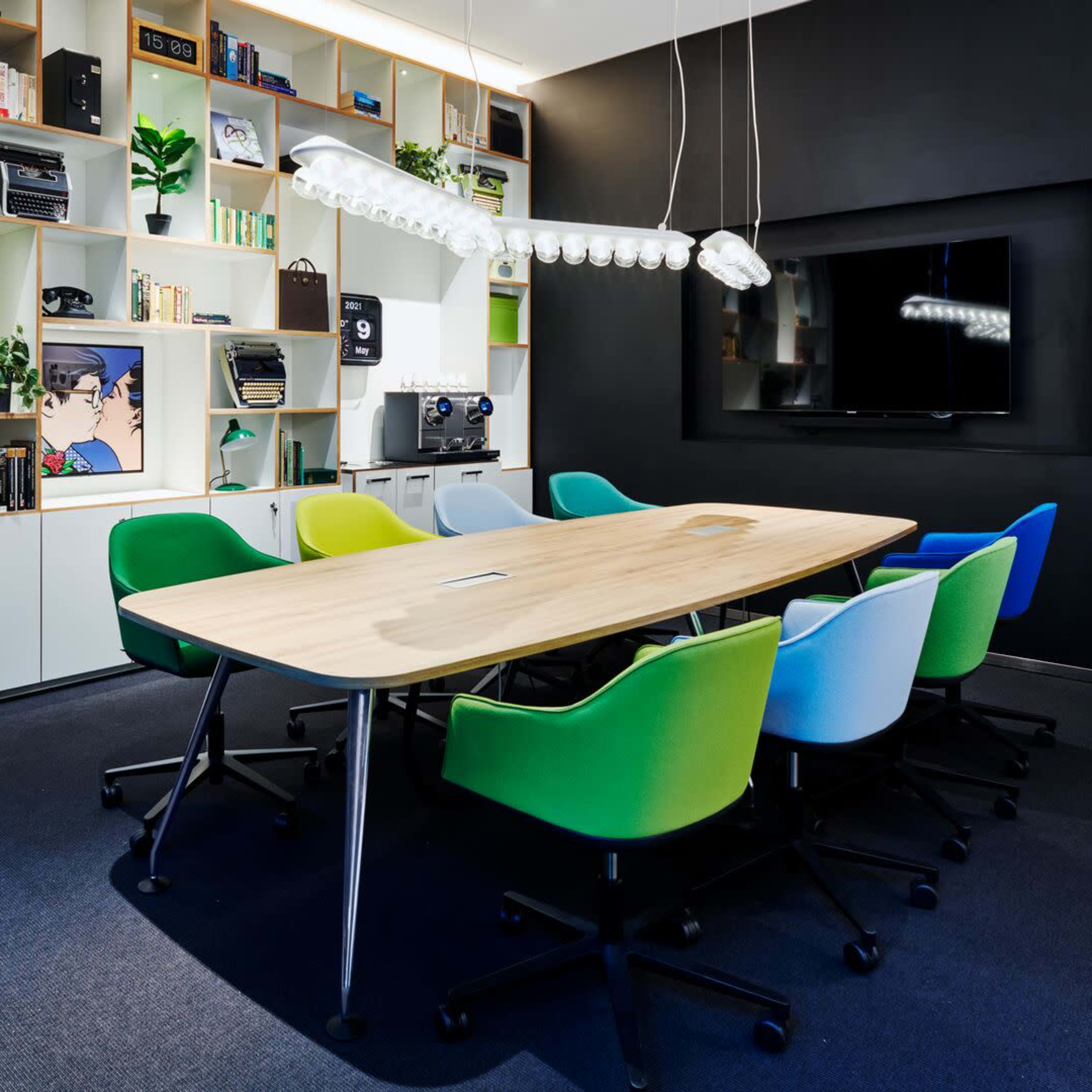 A modern conference room with a long wooden table surrounded by colorful ergonomic chairs and a large wall-mounted screen.