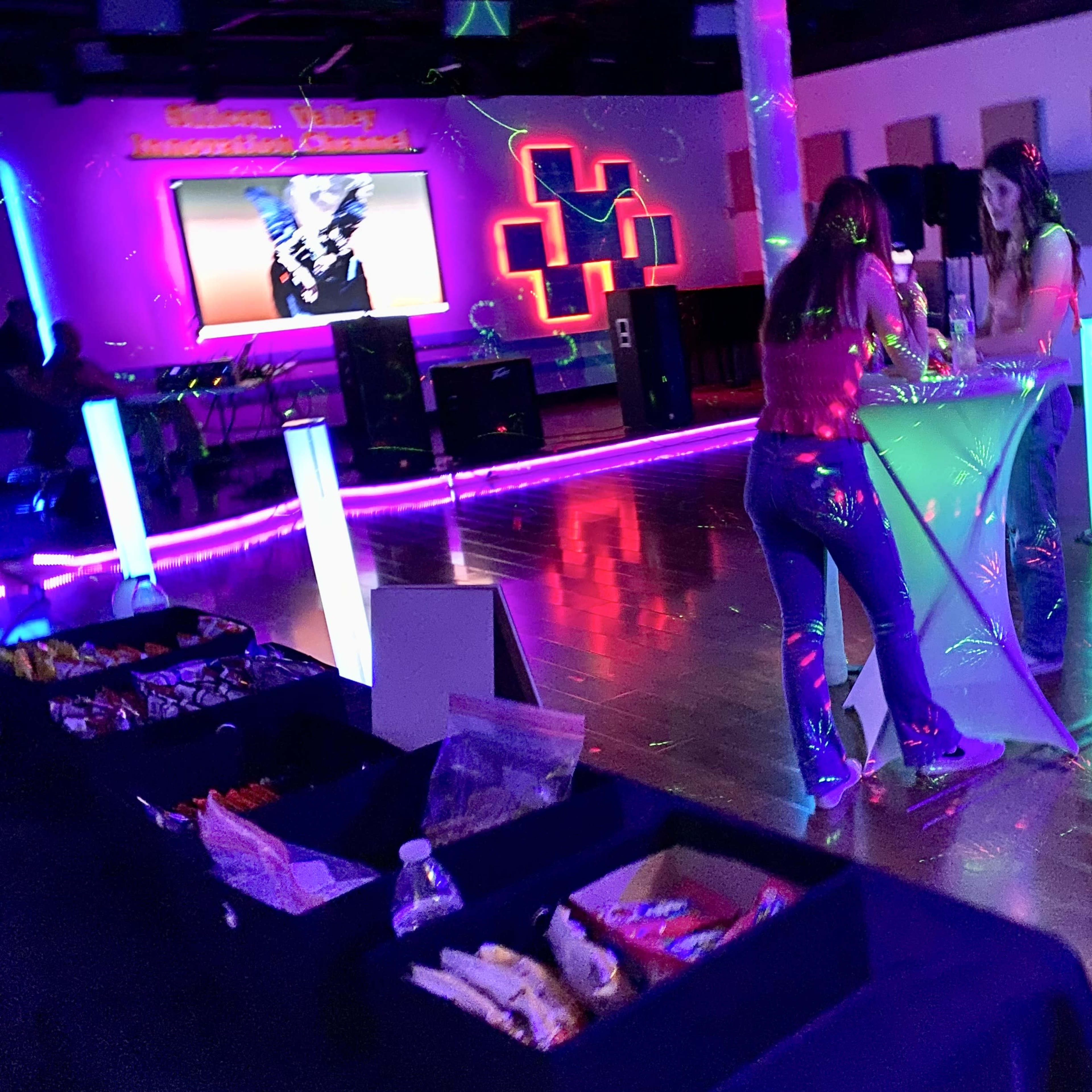 A brightly lit indoor event space with two women engaging at a table, colorful lights reflecting off the walls, and a screen displaying an image in the background.