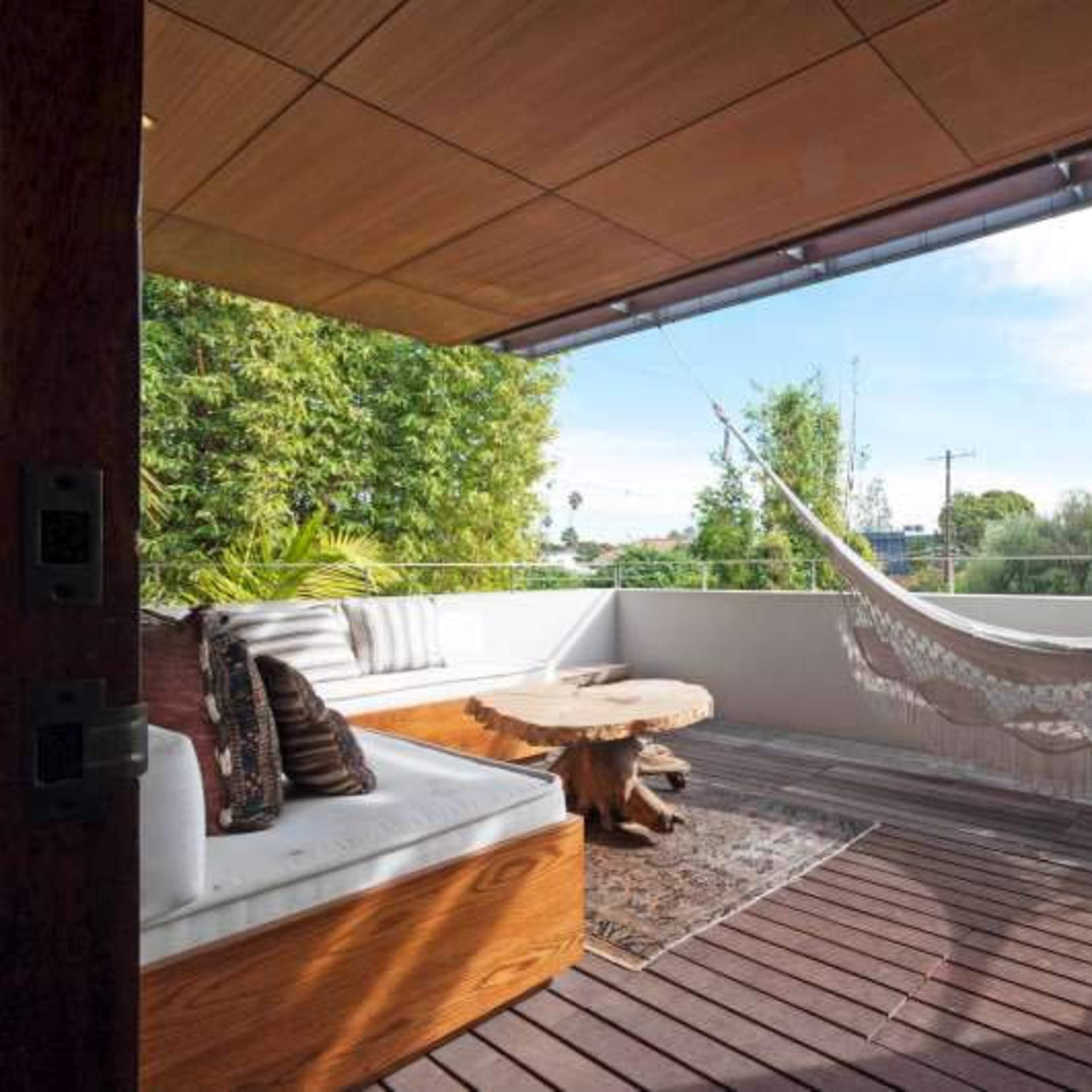 A wooden deck with a hammock, a round wooden table, and seating, surrounded by greenery and open to a blue sky.