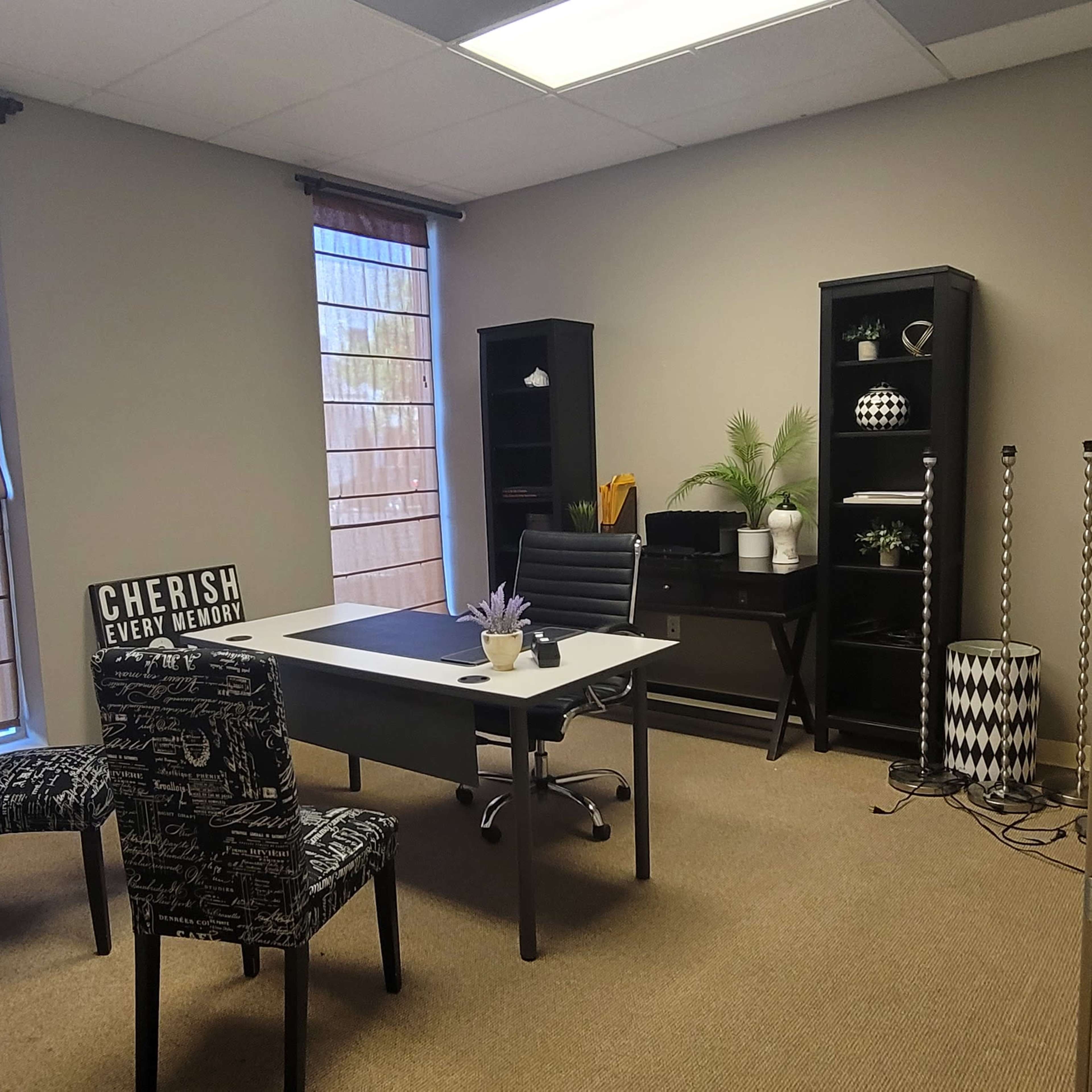 An office space features a desk with a chair, two patterned chairs, a bookshelf, and large windows allowing natural light.