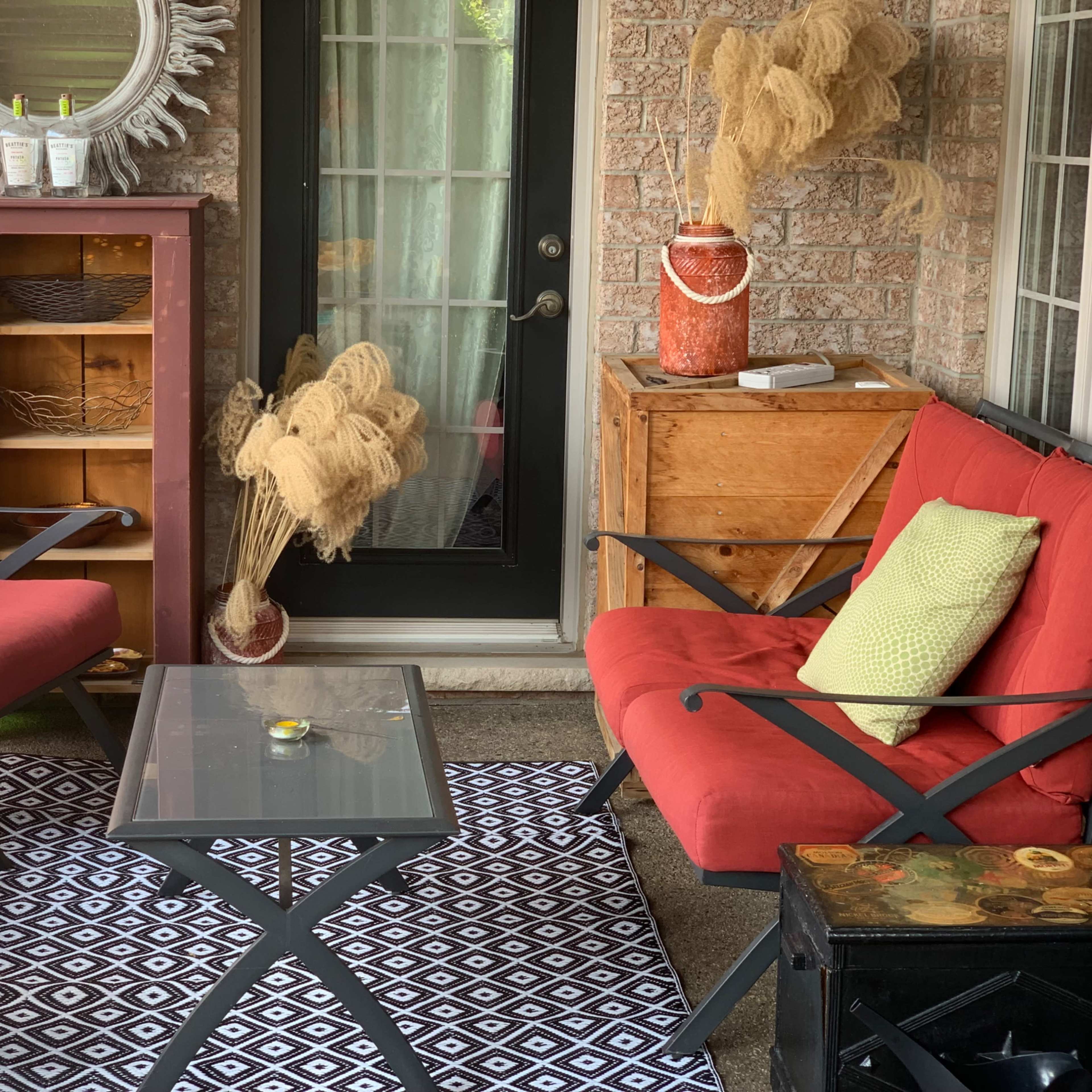 A cozy porch area featuring two red chairs, a glass table, a patterned rug, and decorative elements like a mirror and potted plants.