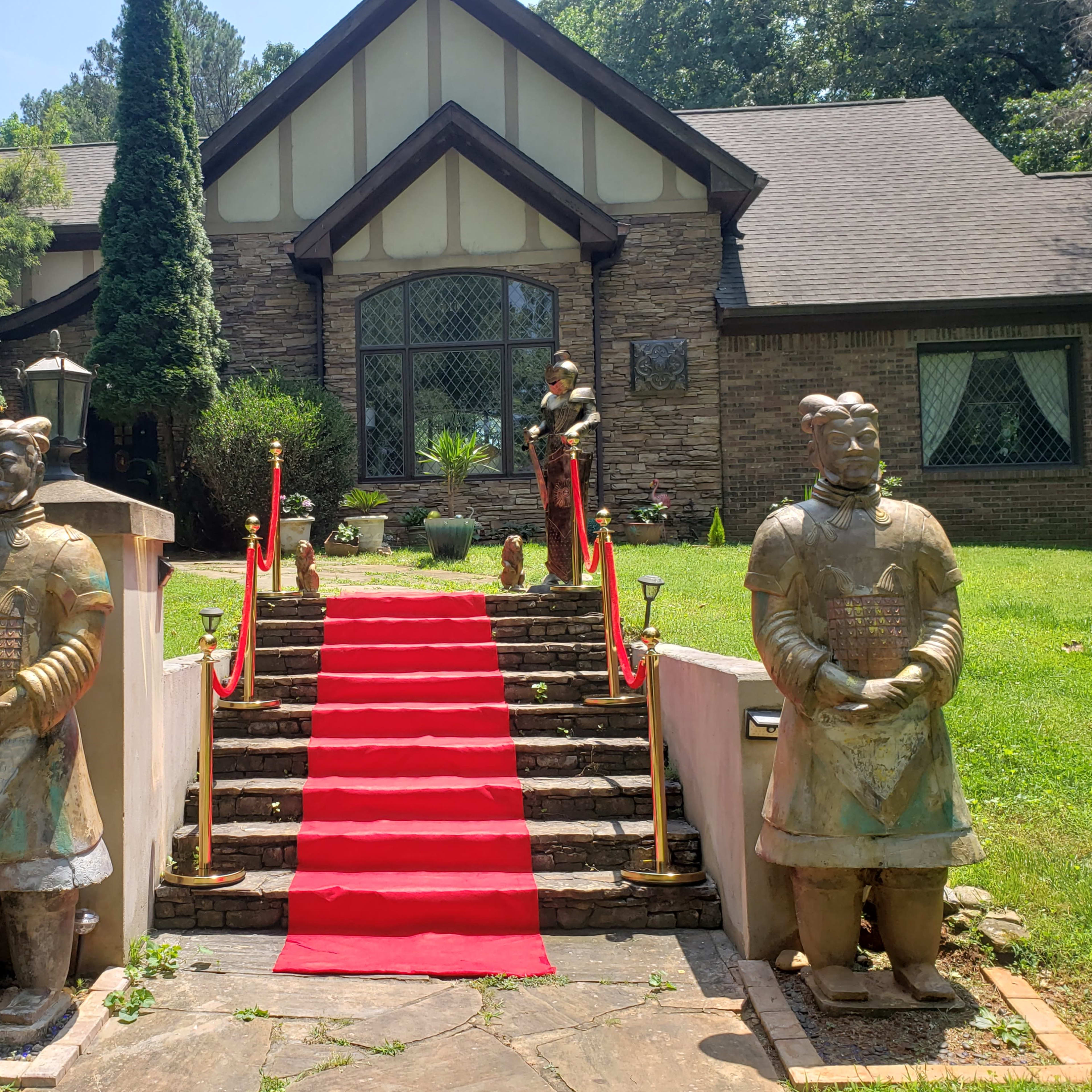 A pathway lined with red carpet leading up to a large stone house, flanked by two statue-like figures resembling ancient warriors.