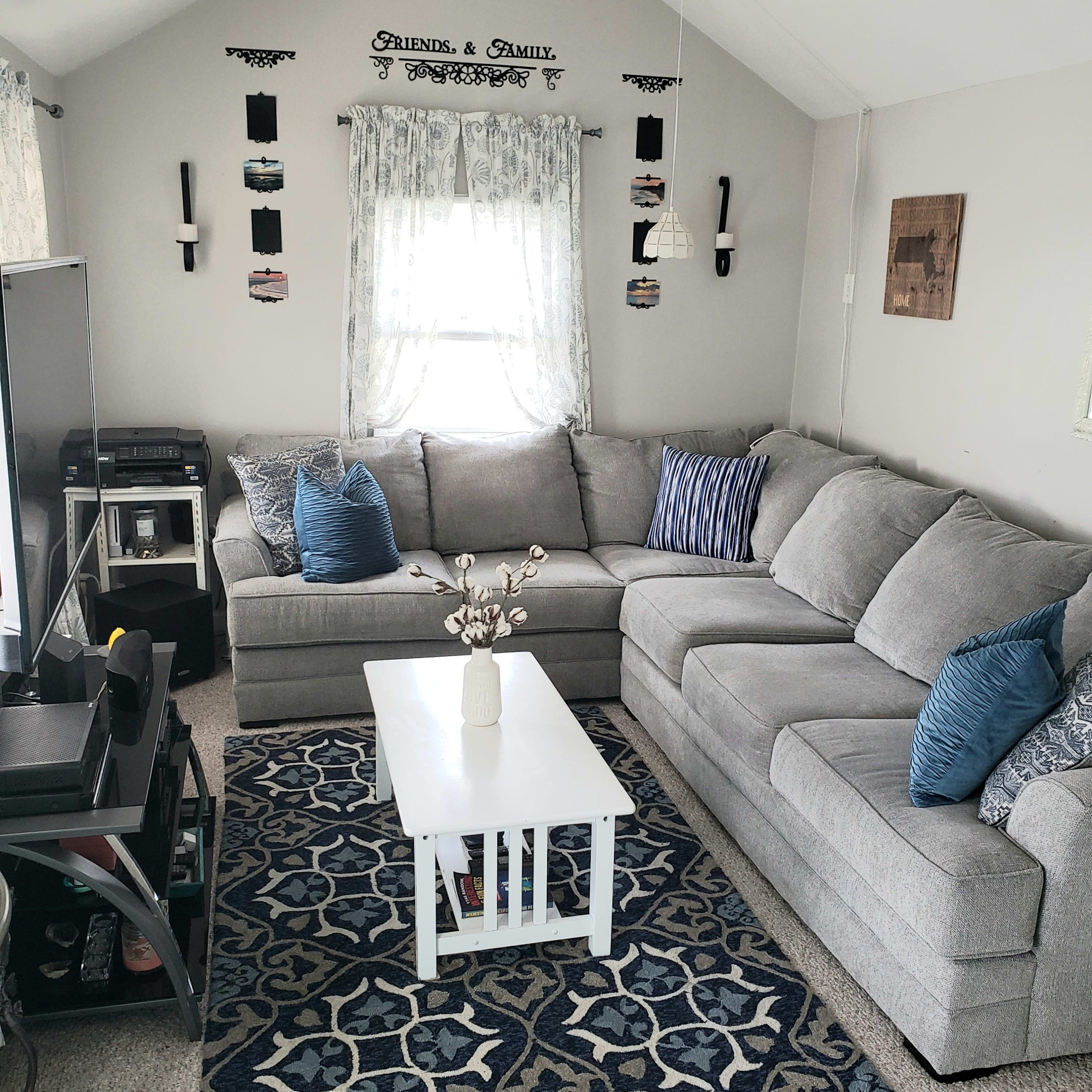 The living room features a grey sectional sofa, a white coffee table, and a patterned blue rug, with windows adorned by light curtains.