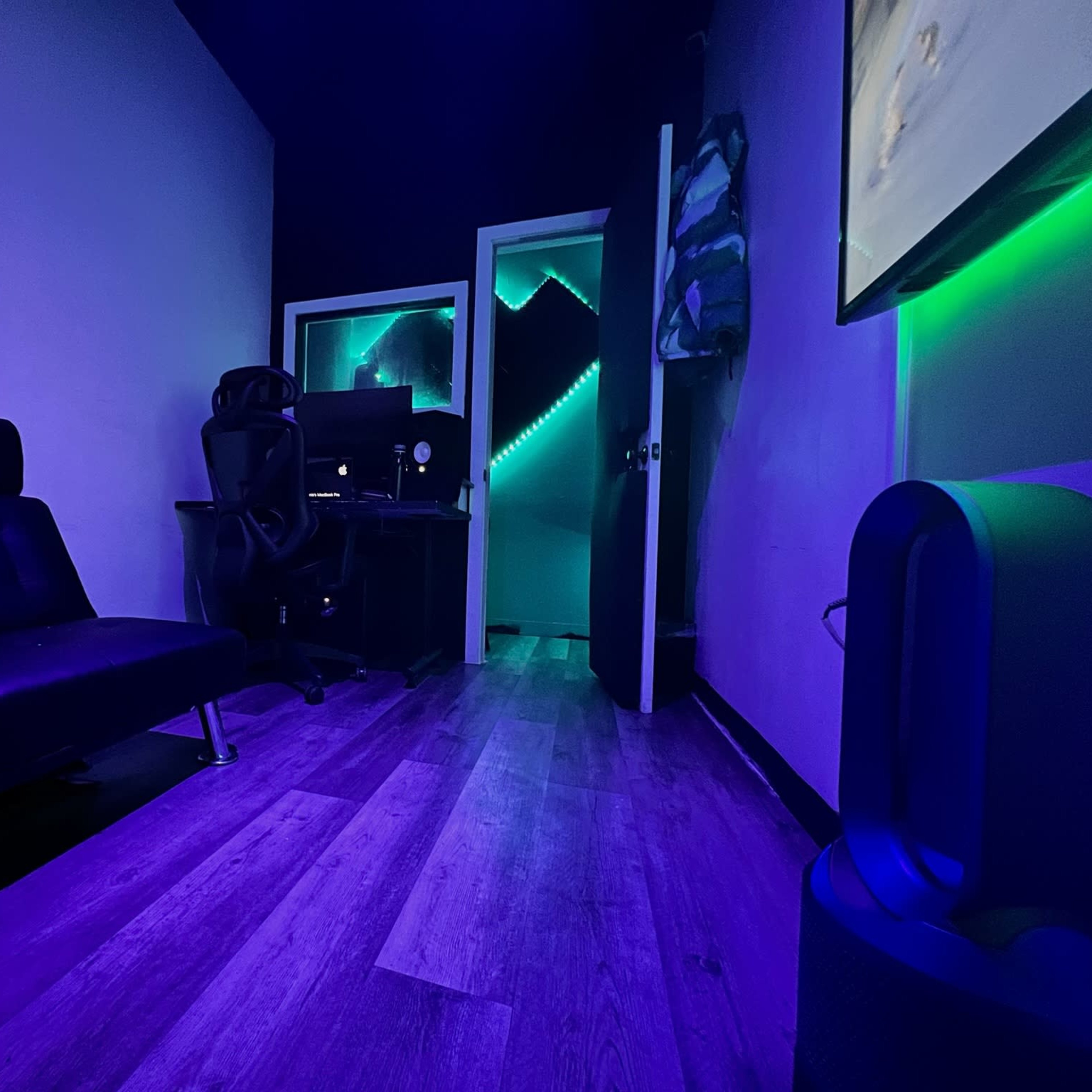 A dimly lit room with a black sofa, a desk with a computer chair, and neon lighting leading to an adjacent room.